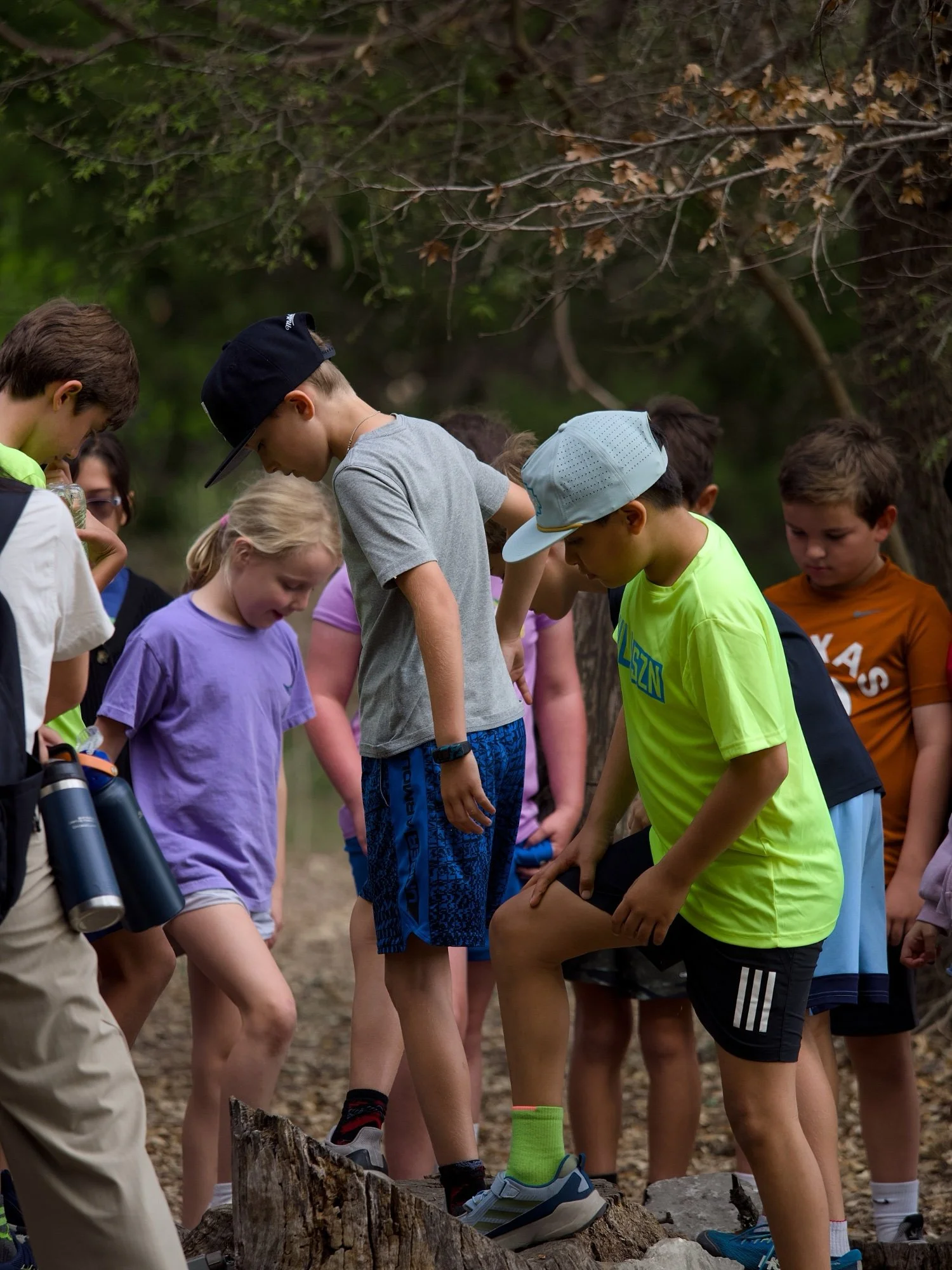 A group of children and teenagers hiking outdoors, with some standing on a large fallen tree log and others standing nearby, surrounded by trees and natural scenery.