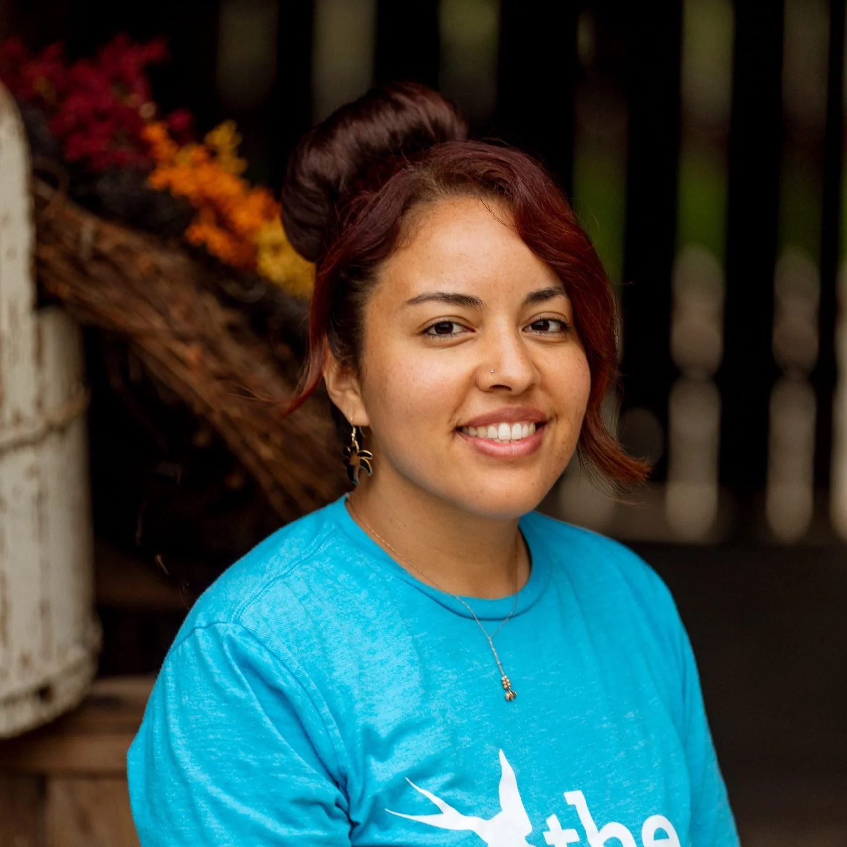 Close-up of a young woman with reddish-brown hair styled in an updo, smiling, wearing a bright blue shirt, with a background featuring orange and red flowers and dark vertical wooden slats.