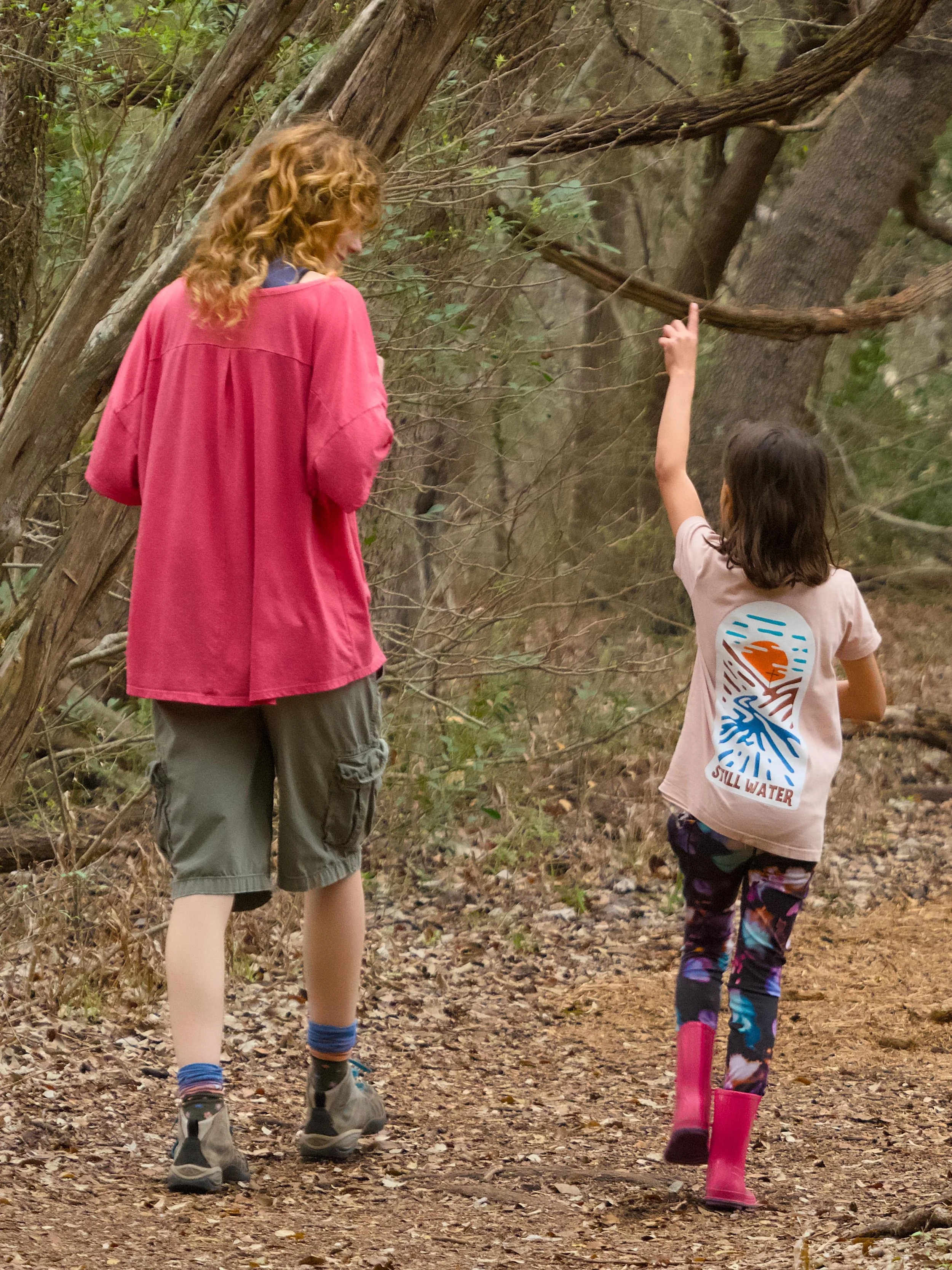 A woman and a girl walking on a trail in a wooded area. The woman has curly hair, is wearing a pinkish jacket, cargo shorts, and hiking boots. The girl has straight hair, is wearing a t-shirt with a colorful graphic, patterned leggings, and pink rain