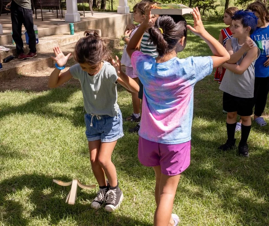 Children playing on a grassy lawn, with one girl balancing a book on top of her head.