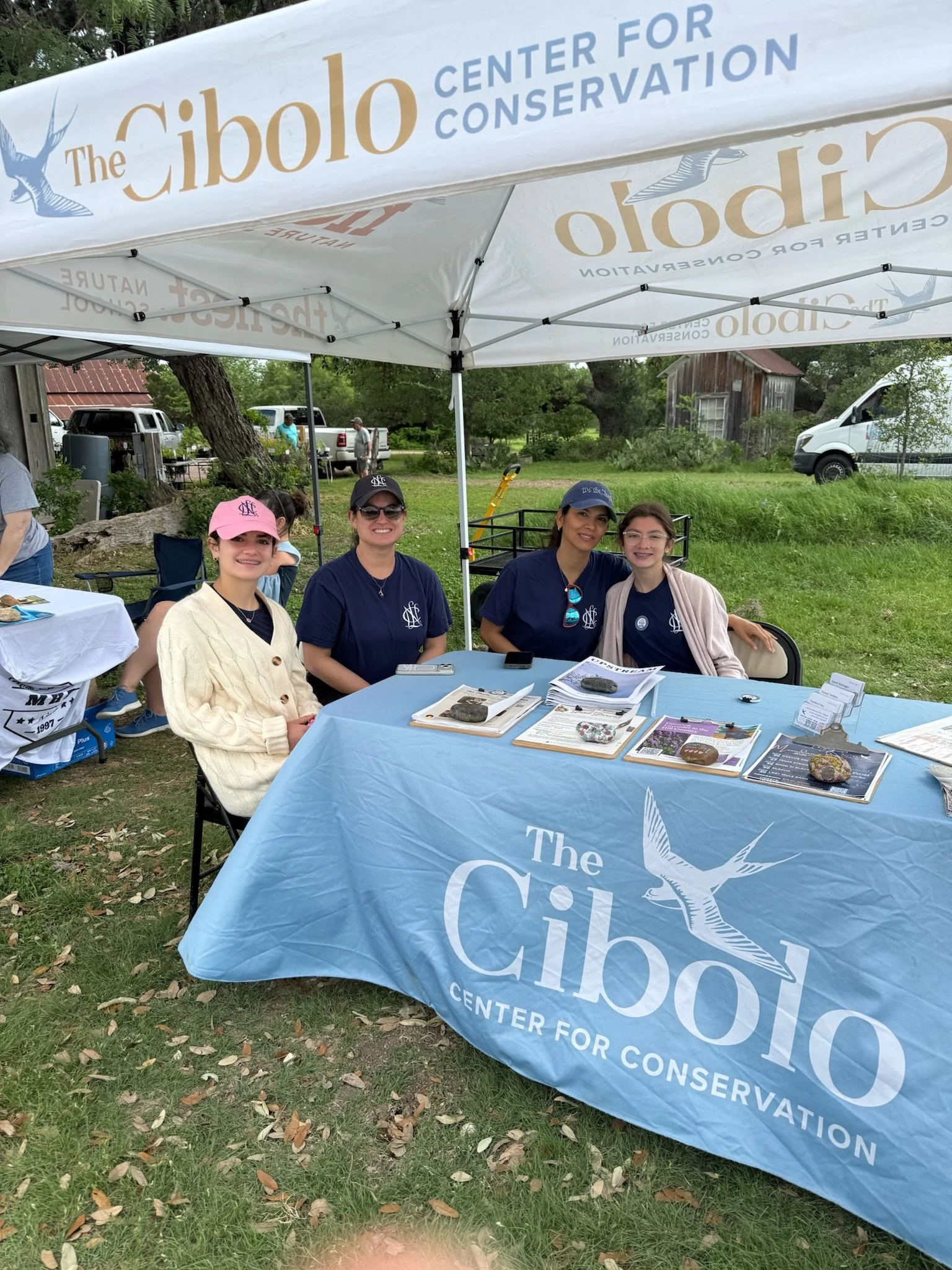 A group of four women sitting under a white canopy tent with blue and gold lettering that reads 'The Cibolo Center for Conservation.' They are outdoors with a grassy area, trees, and a rustic building in the background. The women are smiling and seat