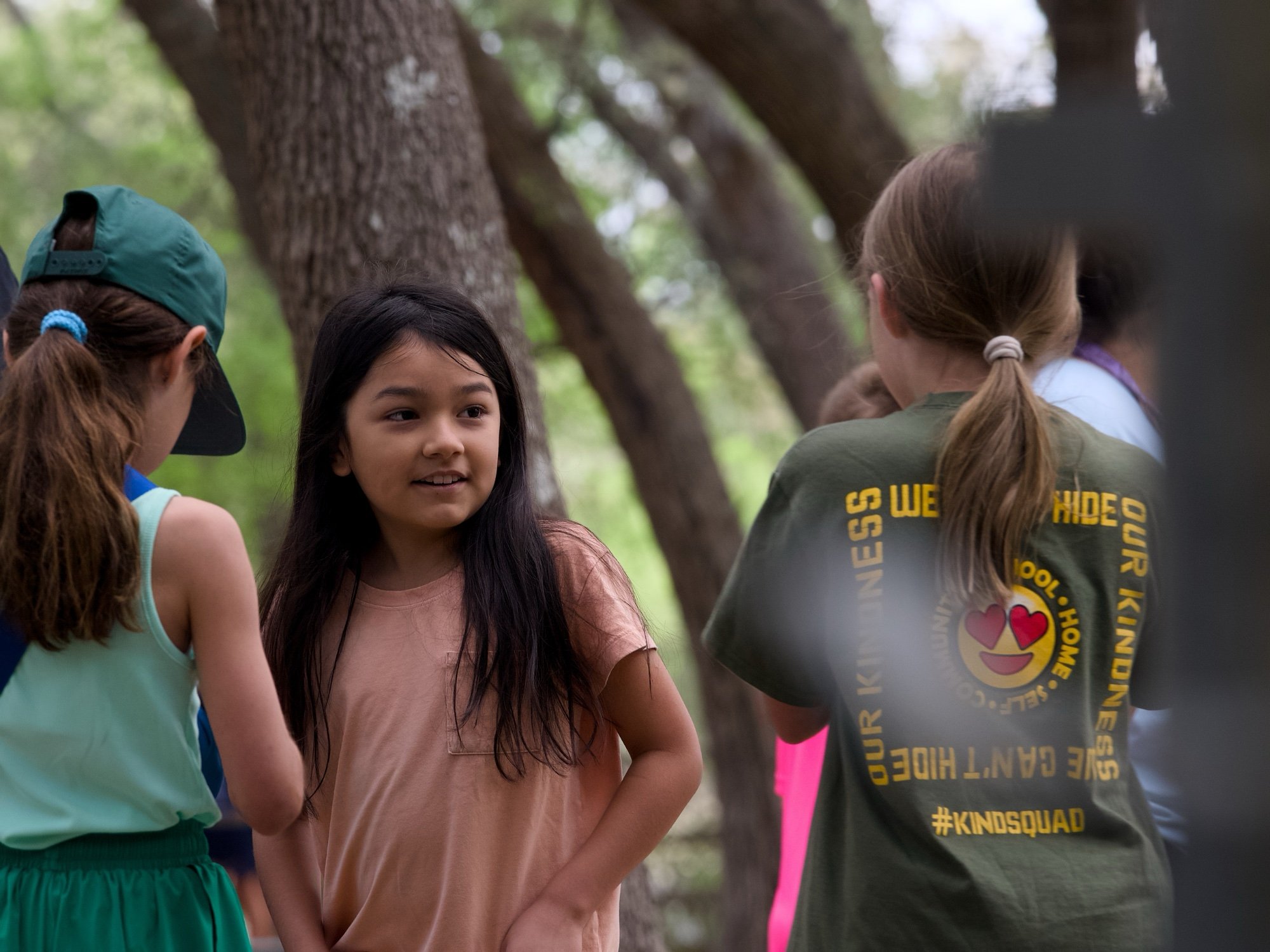 A group of children standing outdoors in a wooded area, engaged in conversation, with trees and natural surroundings in the background.