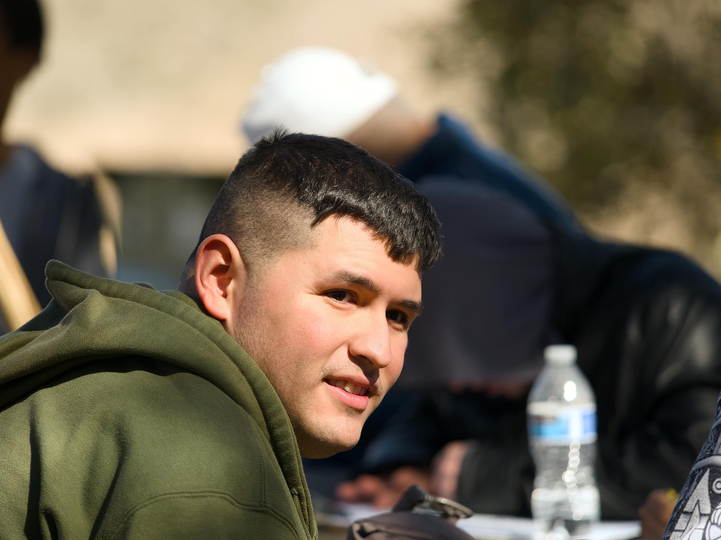 Close-up of a young man with short dark hair, wearing a green hoodie, smiling slightly, with other people and a water bottle in the background.