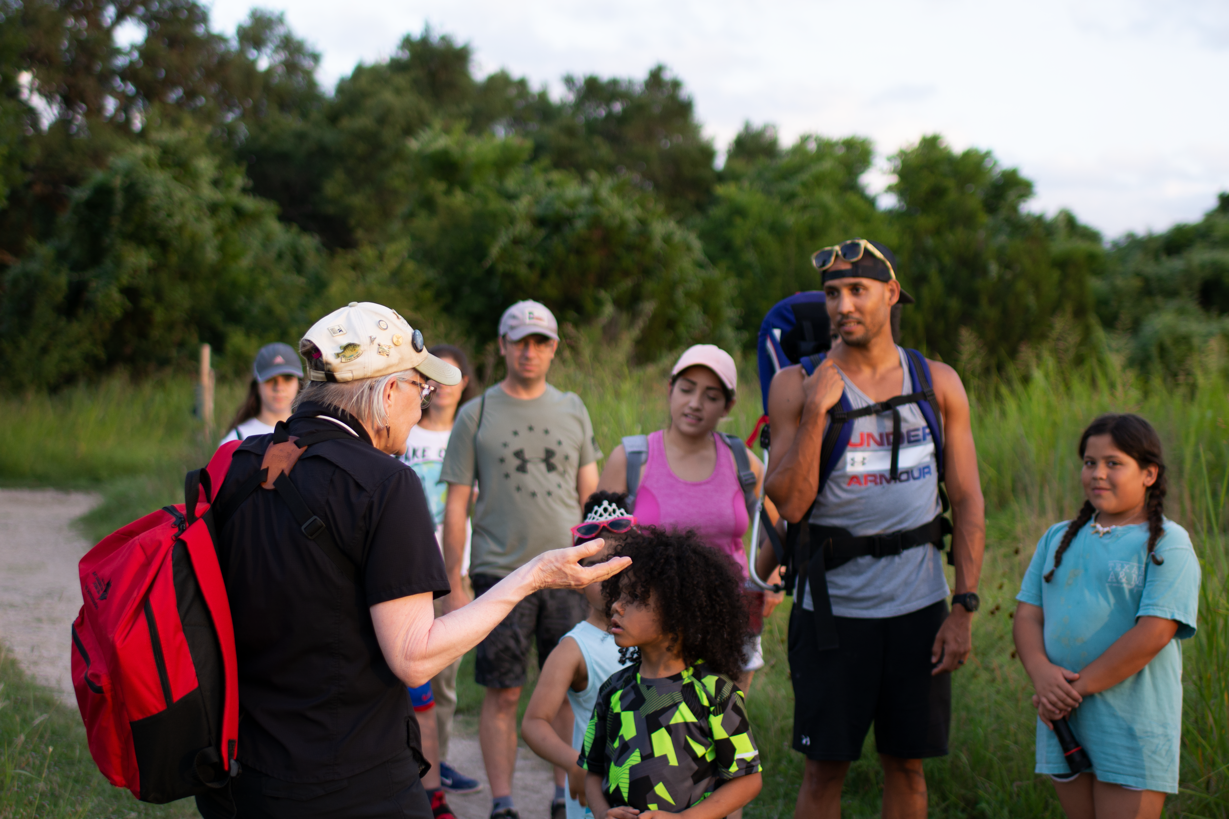 Group of children and teenagers outdoors on a nature hike talking to an older woman with a red backpack and a beige cap, surrounded by green trees.