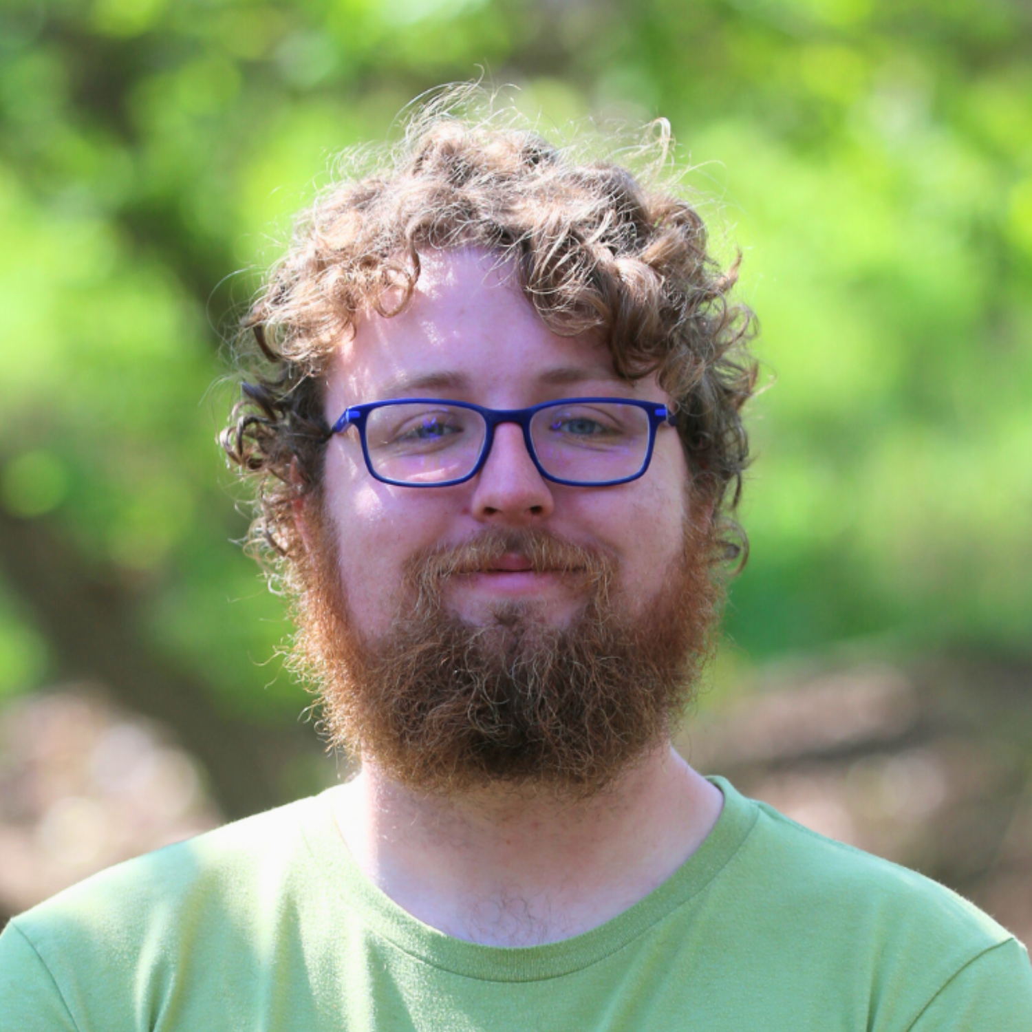 Portrait of a man with curly hair and glasses, smiling, outdoors with blurred green foliage in the background.