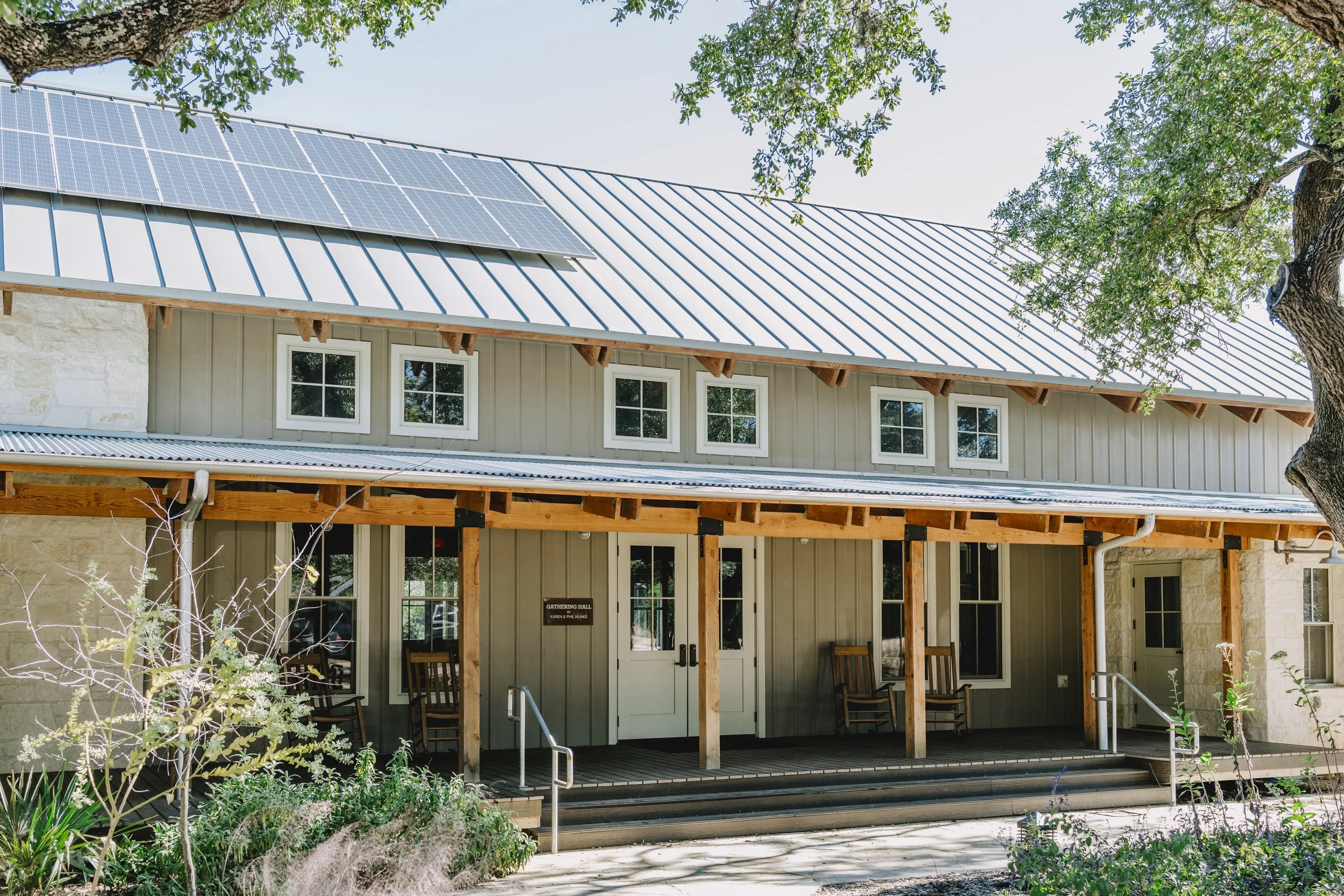 The image shows the exterior of a building with a metal roof, which has solar panels installed on the left side. There are multiple windows on the upper floor and a door below, with a gathering hall sign beside it. The front area near the entrance ha