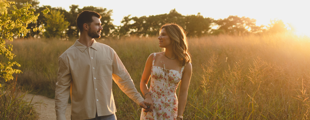 A young couple holding hands walks through a grassy prairie at sunset, with trees in the background.