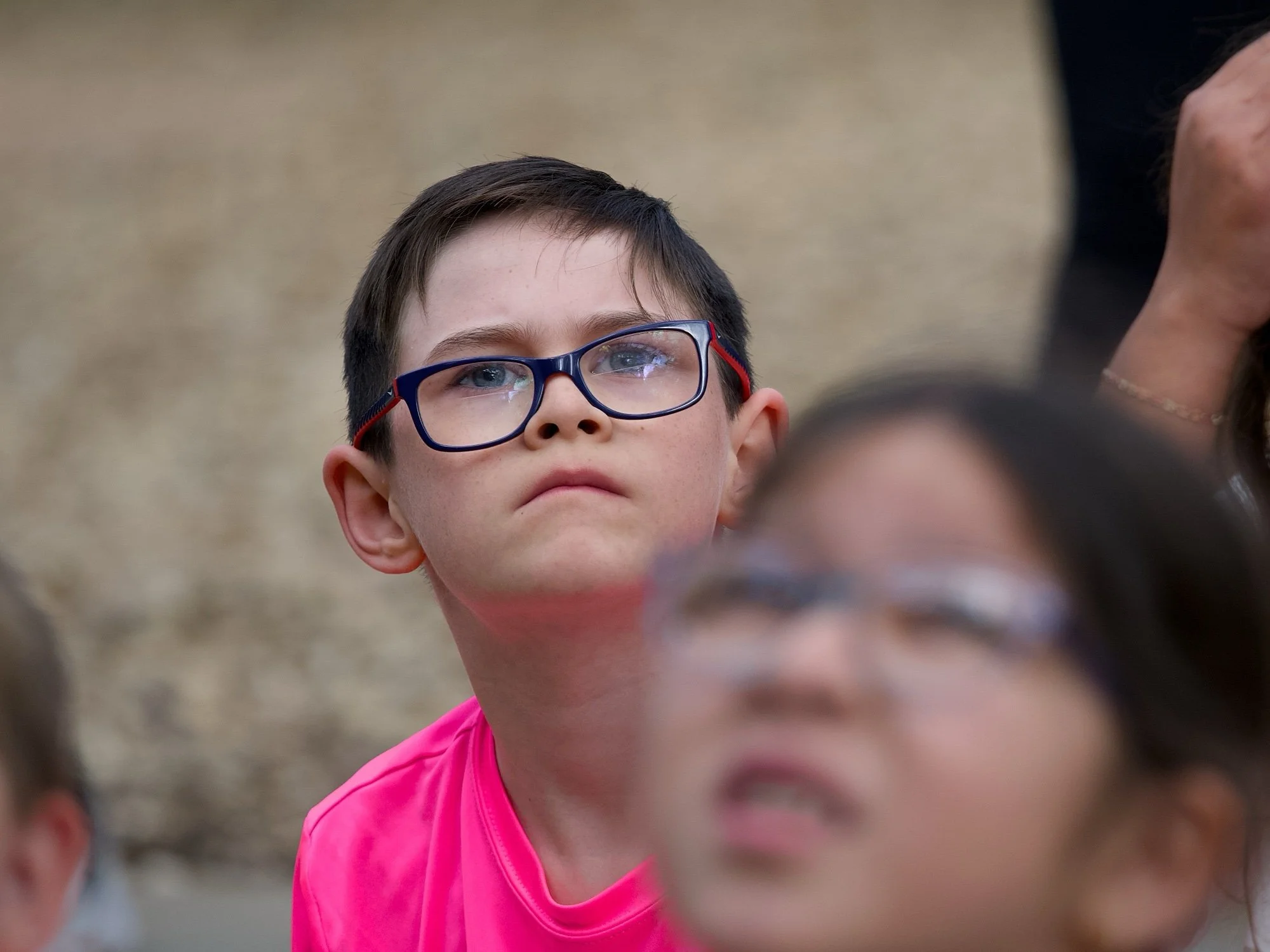 Close-up of a young boy wearing glasses and a pink shirt, with other blurred children and people in the background.