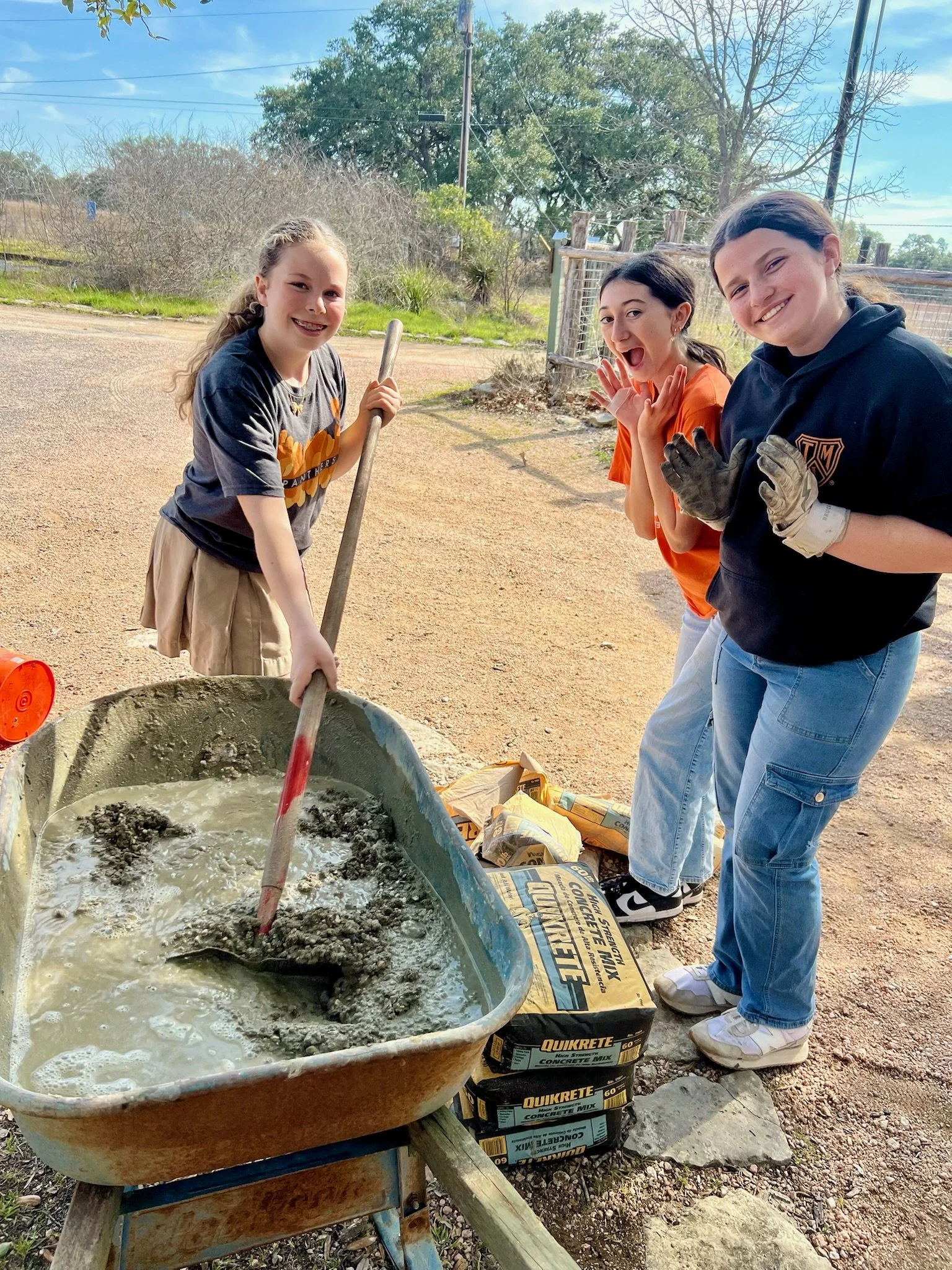 Three young girls are working together outdoors, mixing concrete in a wheelbarrow. They appear happy and engaged in a construction or gardening activity, with bags of concrete mix nearby.