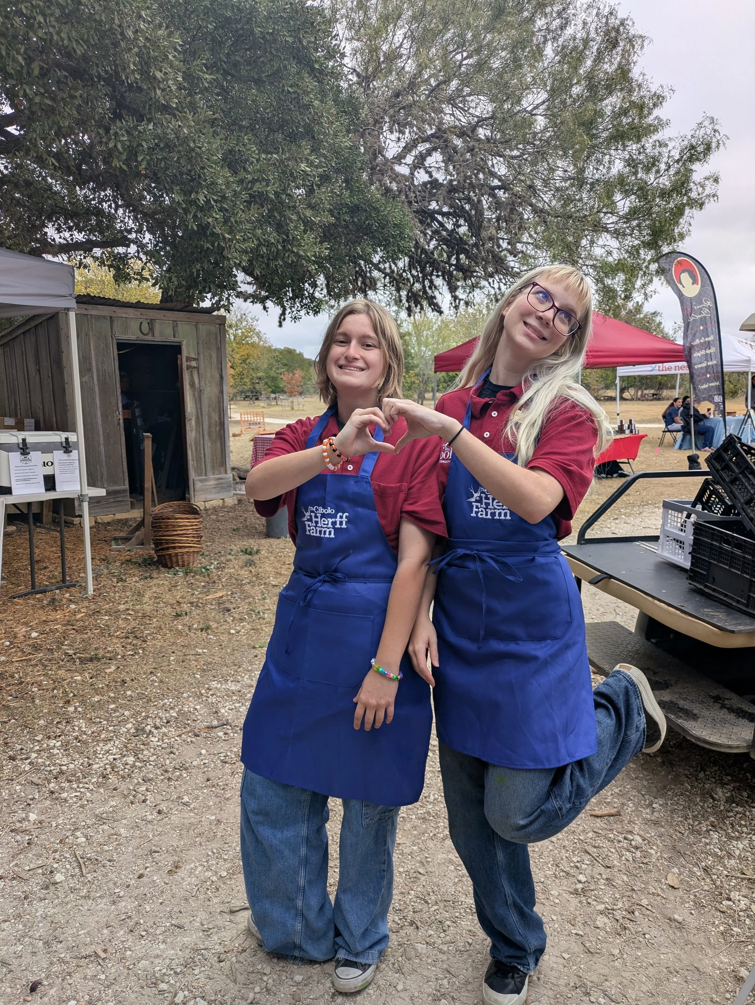 Two smiling young women standing outdoors under trees, making a heart shape with their hands, wearing blue aprons with the text "The Cibolo Herrf Farm," at a community farmers market with tents and people in the background.