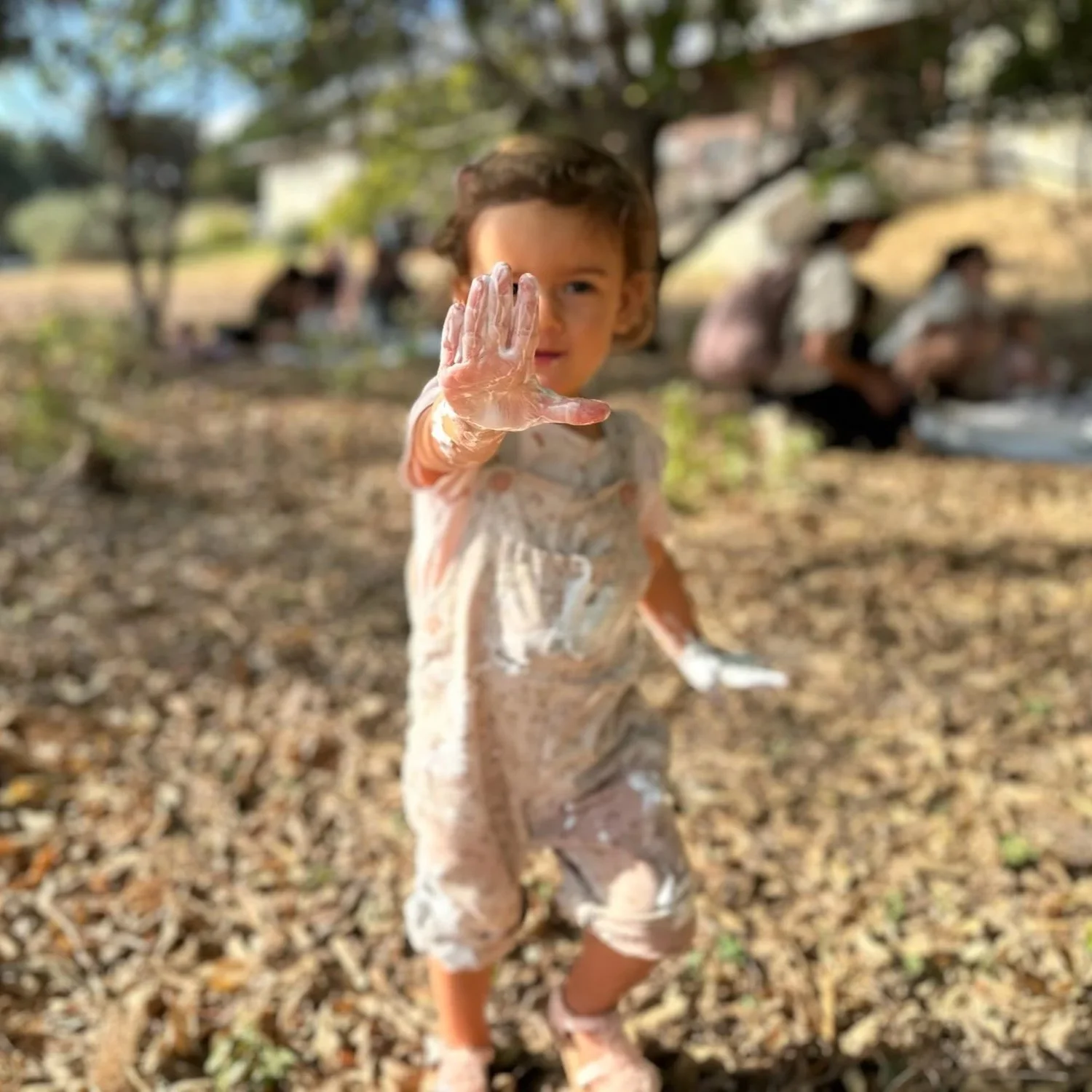 A young girl with messy hair and paint on her face and clothes, raising her hand covered in white paint or chalk, outdoors with trees and blurred people in the background.