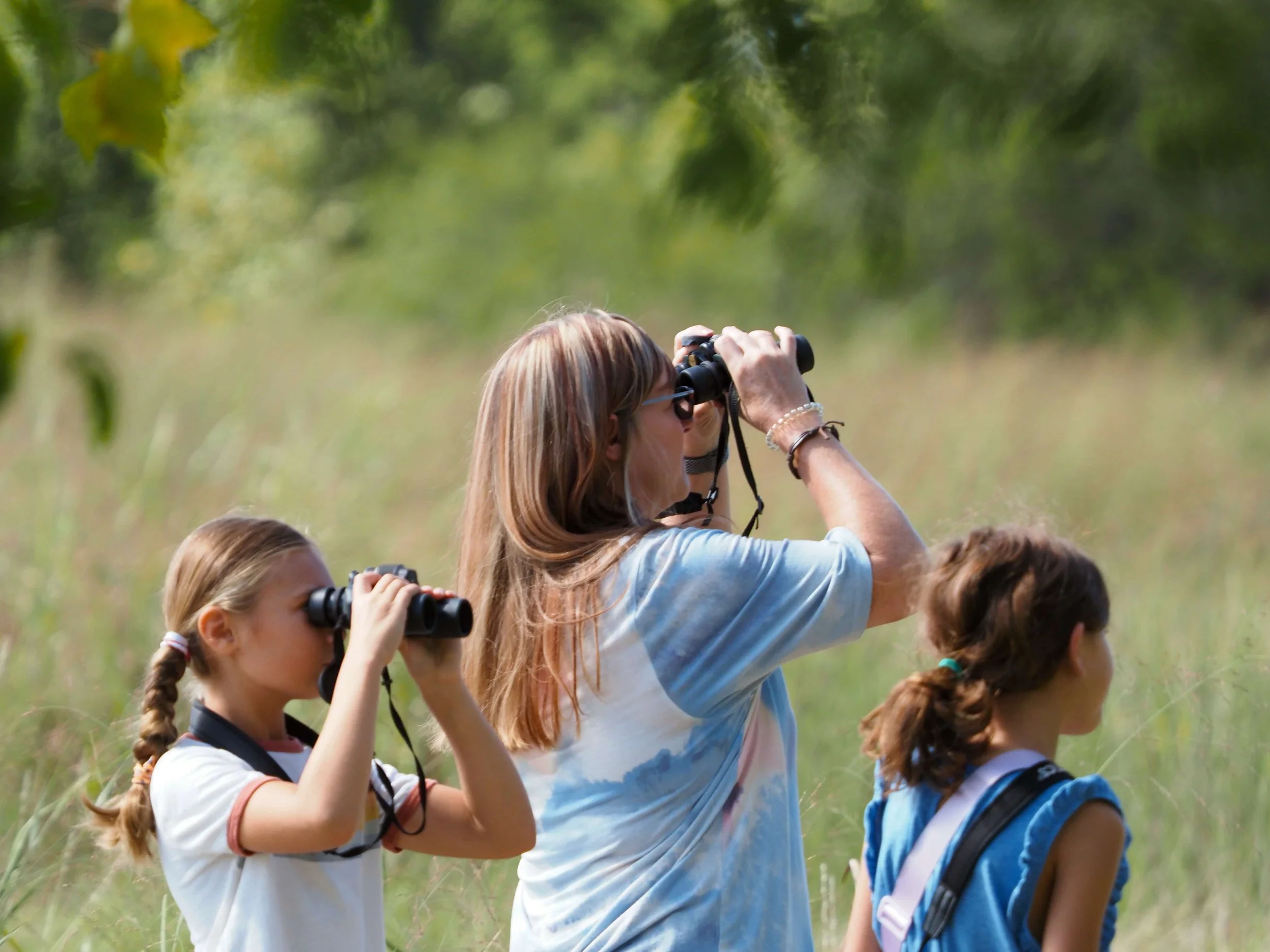 Children and an adult looking through binoculars outdoors in a green grassy native prairie with trees, natural light.