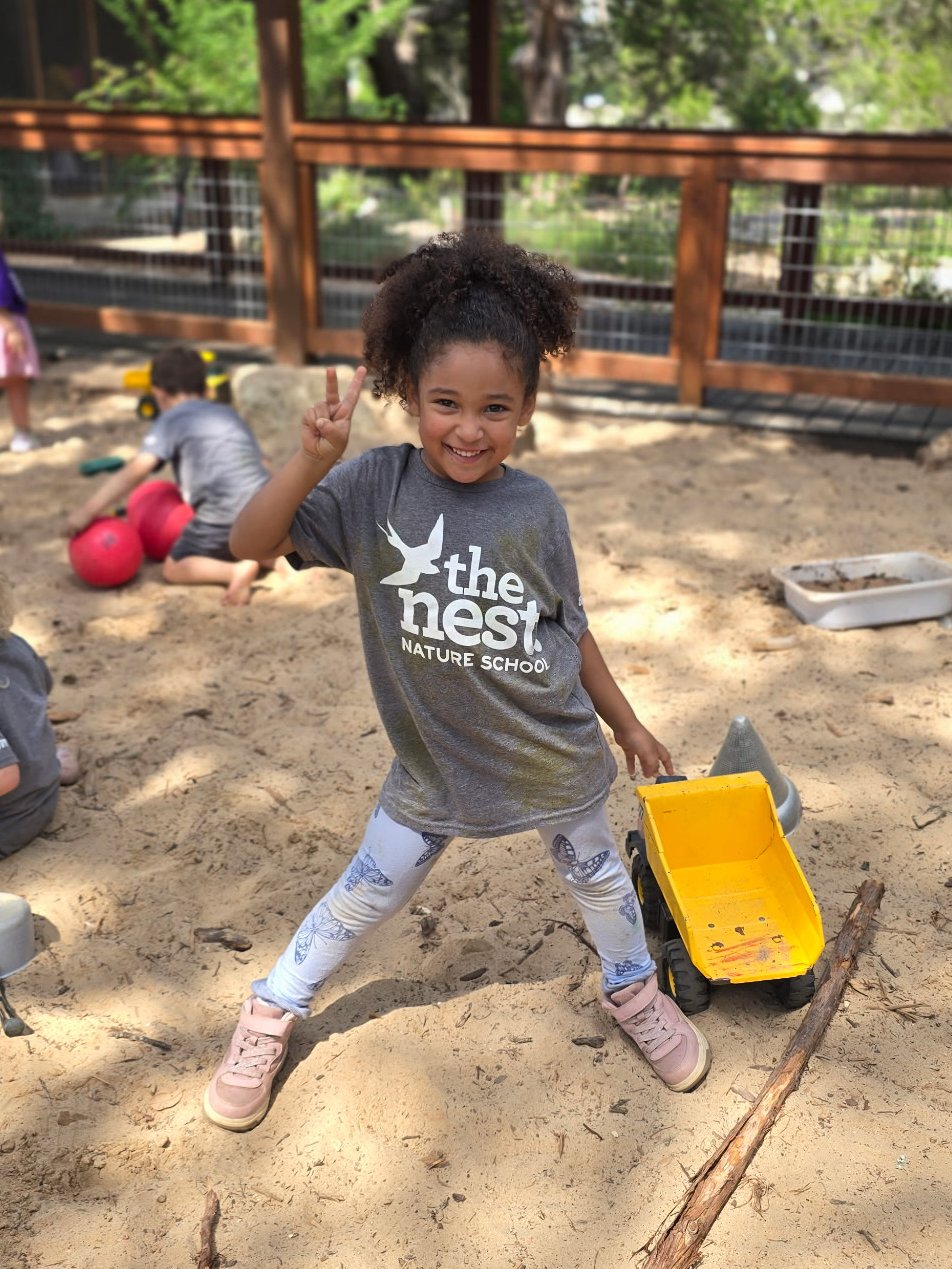 A smiling young girl with curly hair making a peace sign in a sandbox at a playground, wearing a gray T-shirt with the words 'the nest' and 'nature school,' surrounded by sand toys and sticks.