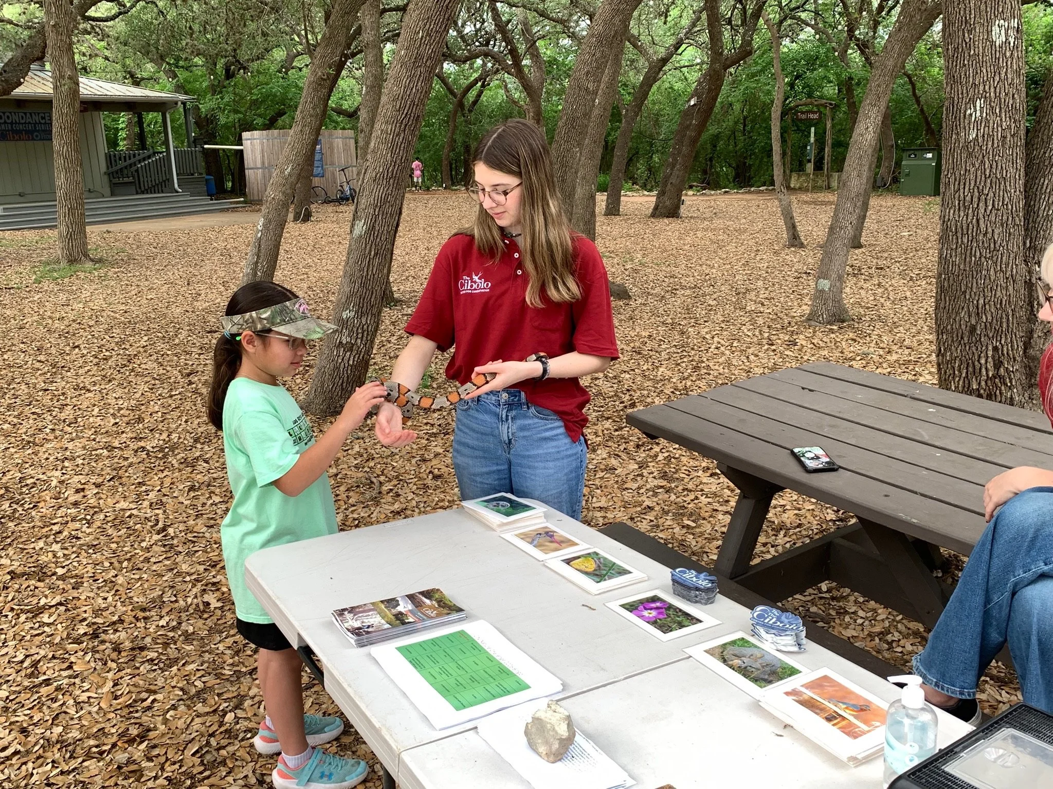 A young girl, an older girl, and another girl are sitting at a table outdoors in a wooded area with fallen leaves on the ground. The young girl has a camouflage cap, glasses, and a light green shirt. The older girl has long hair, glasses, a red shirt