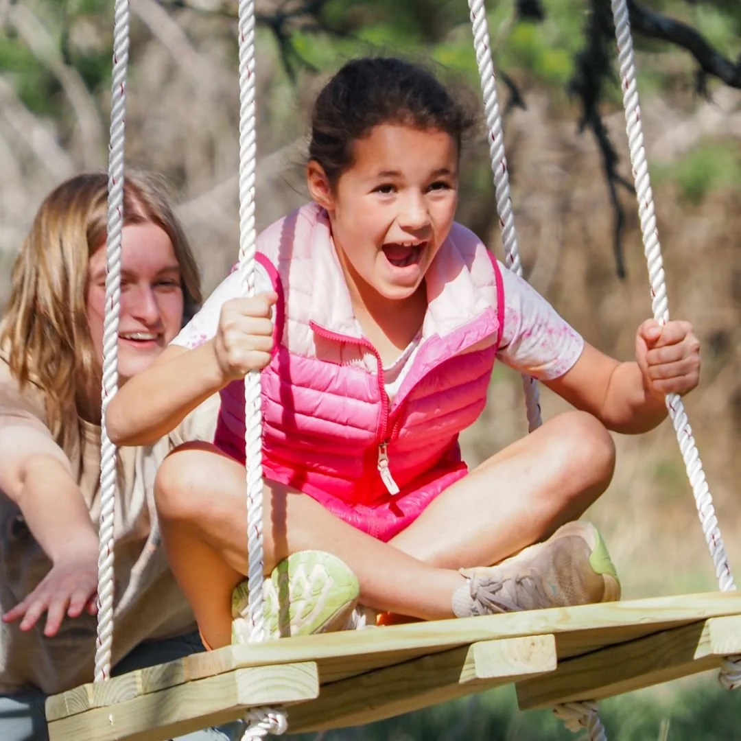 Two girls playing on a wooden swing outdoors, smiling and enjoying a sunny day.
