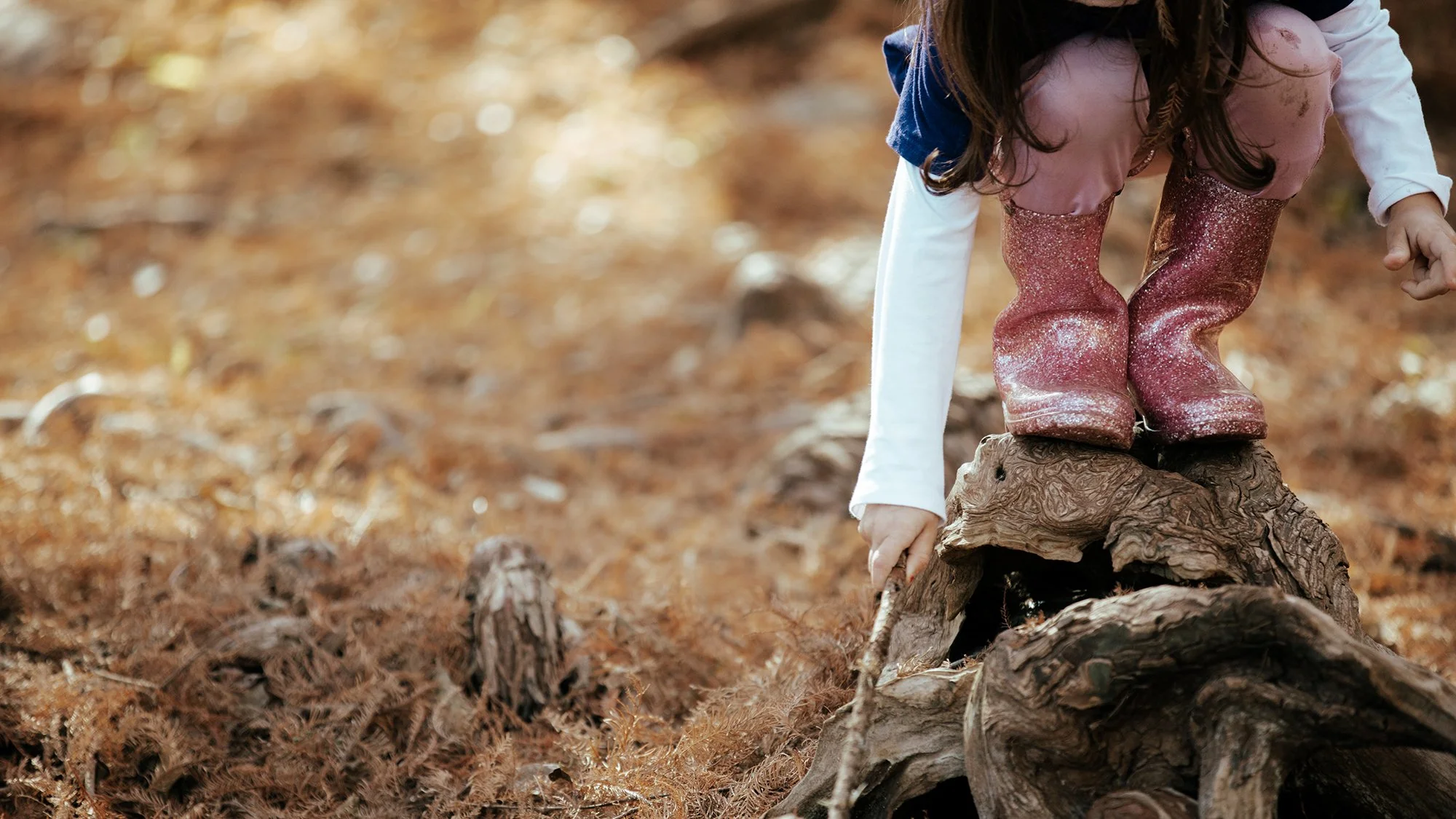 A young girl with long dark hair wearing pink glittery rain boots, white pants, and a pink jacket is crouched on a log outdoors during autumn. She is reaching down with her right hand to touch the ground, surrounded by fallen leaves and small logs.