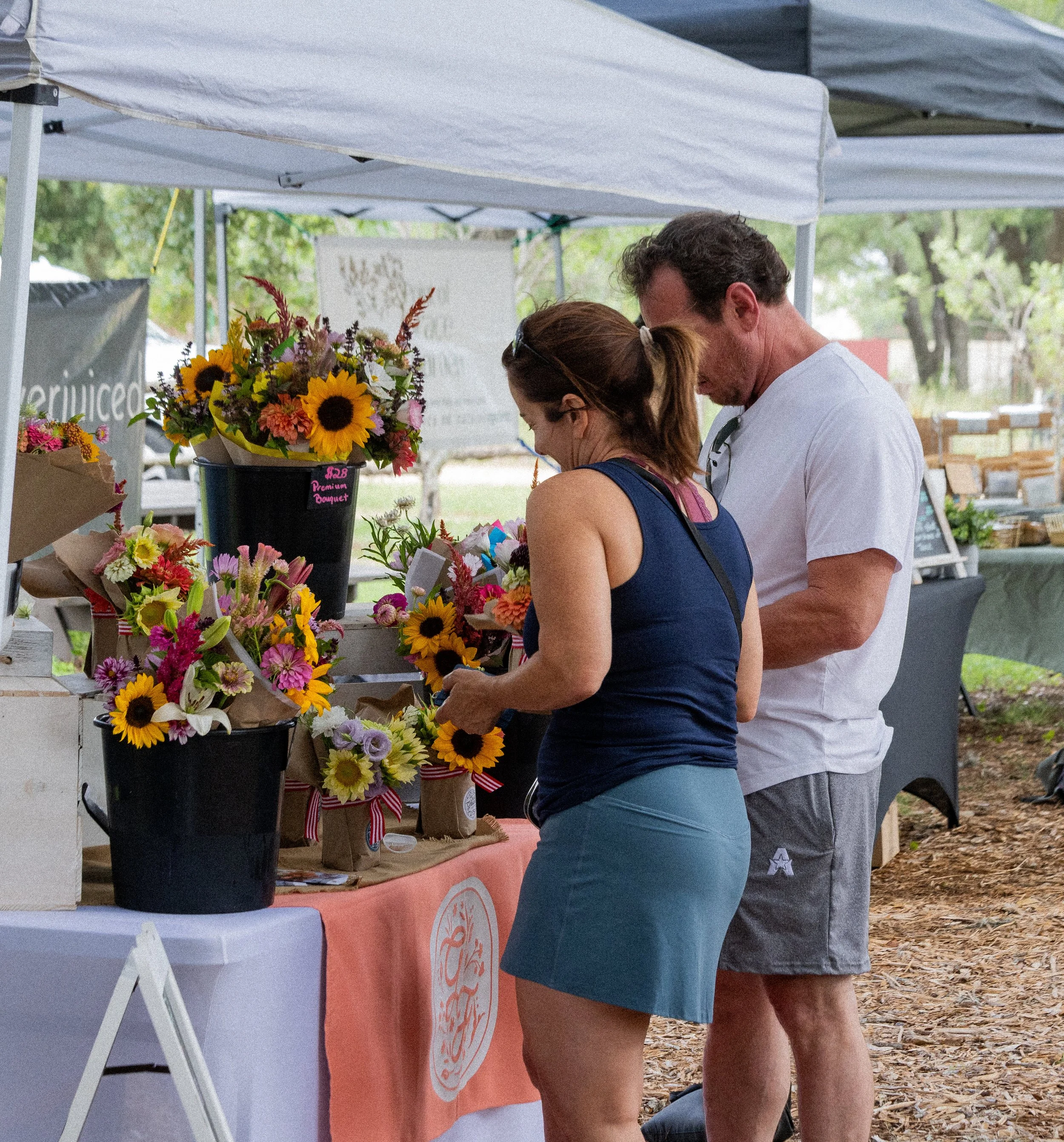 People shopping for flowers at an outdoor market stall under a canopy, with various bouquets and potted flowers displayed.
