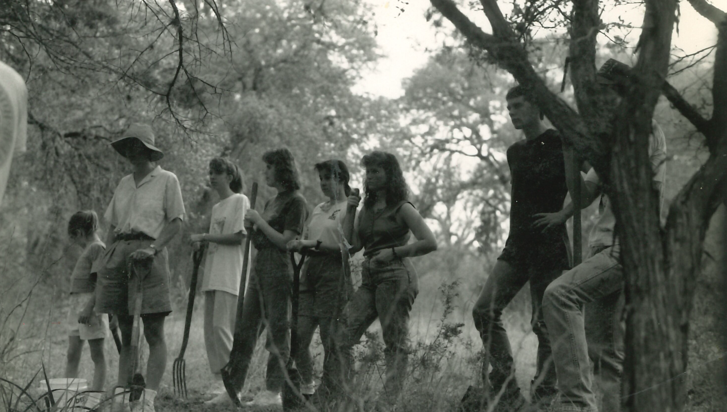 A black and white photo of a group of people standing outdoors among trees, some holding shovels, and a woman speaking into a microphone.