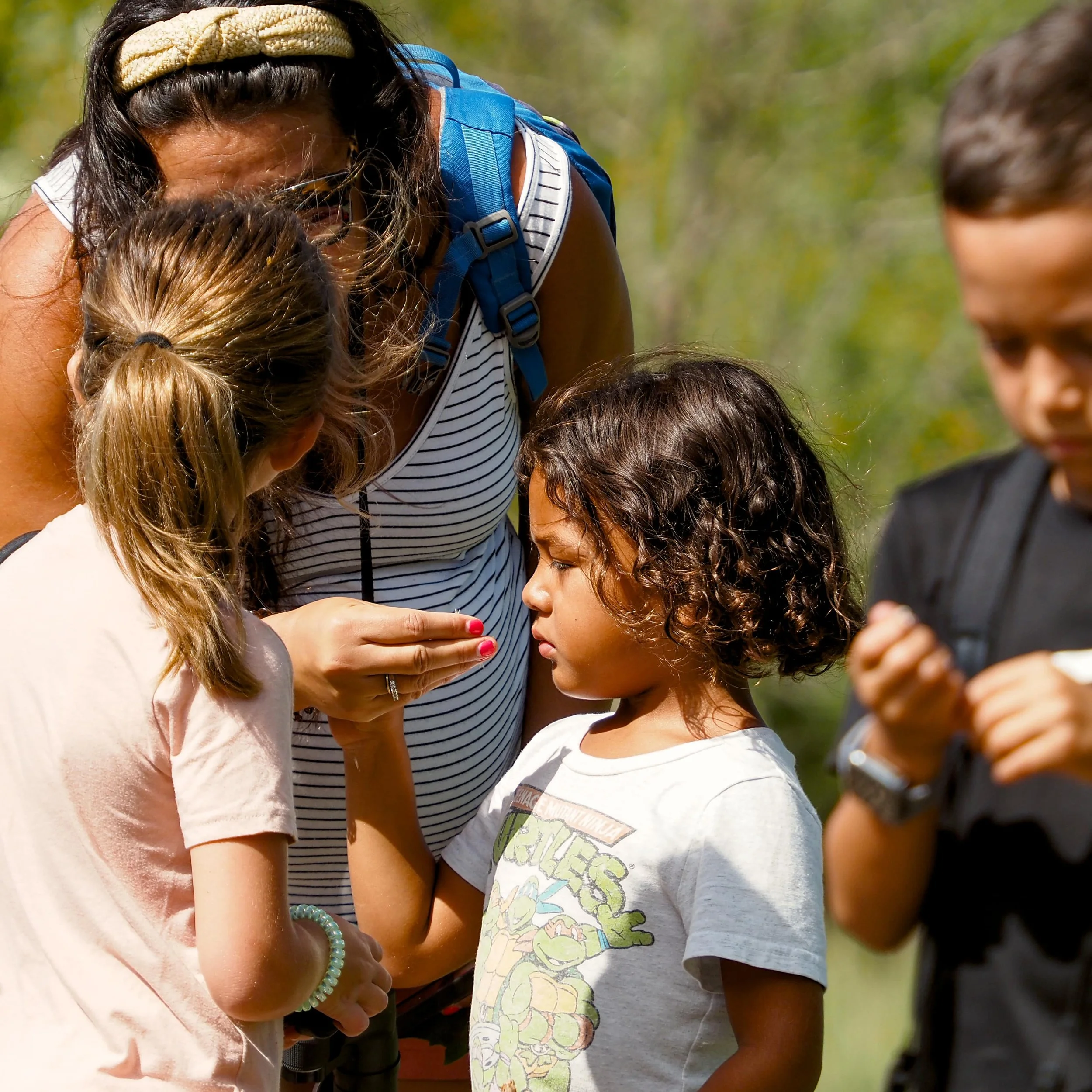 Group of children and an adult closely examining something outdoors, with a woman with dark hair, a headband, and a striped shirt.
