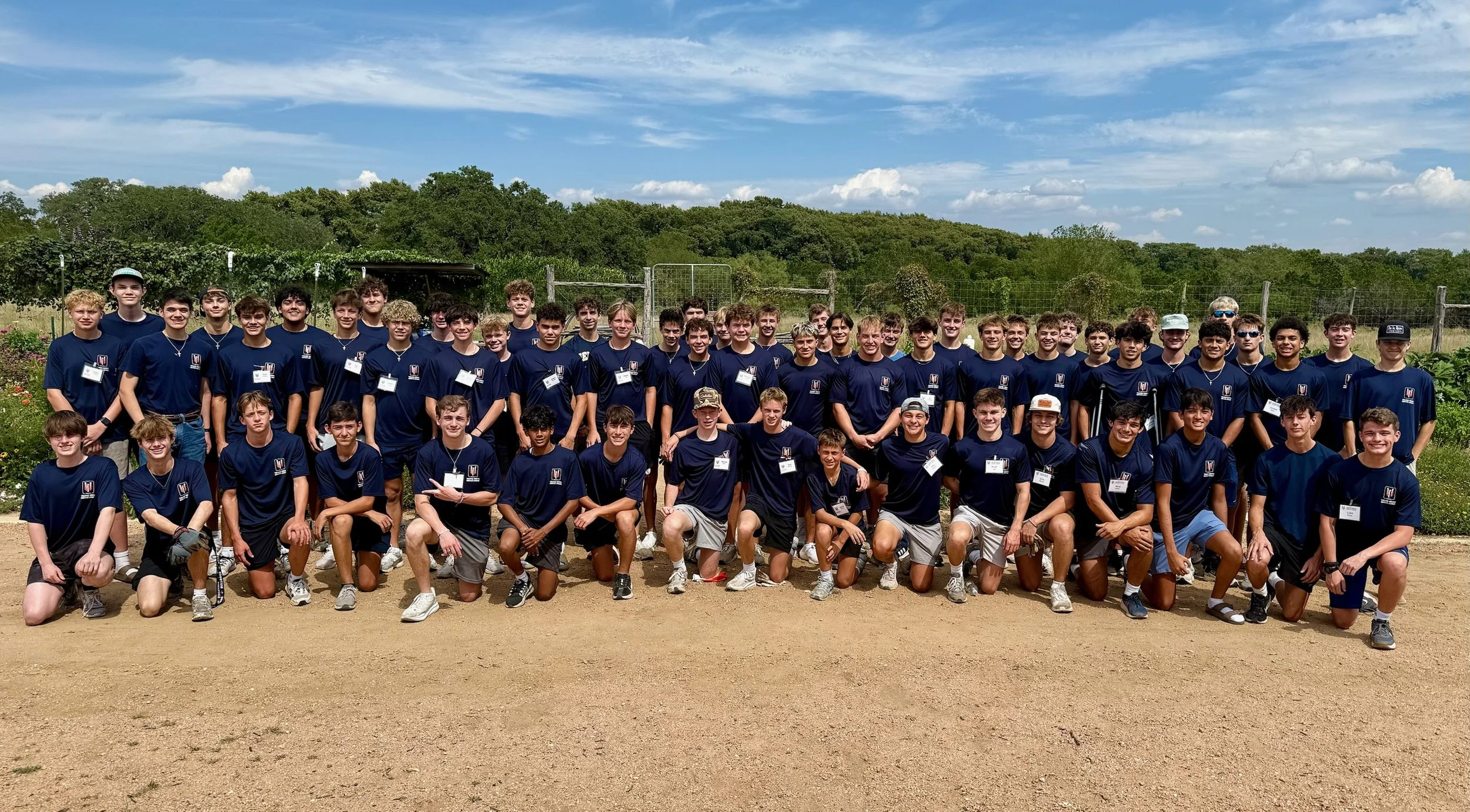 Group of young boys and teenagers posing outdoors on a dirt field, wearing matching navy blue T-shirts with a logo, under a partly cloudy sky, with green trees in the background.