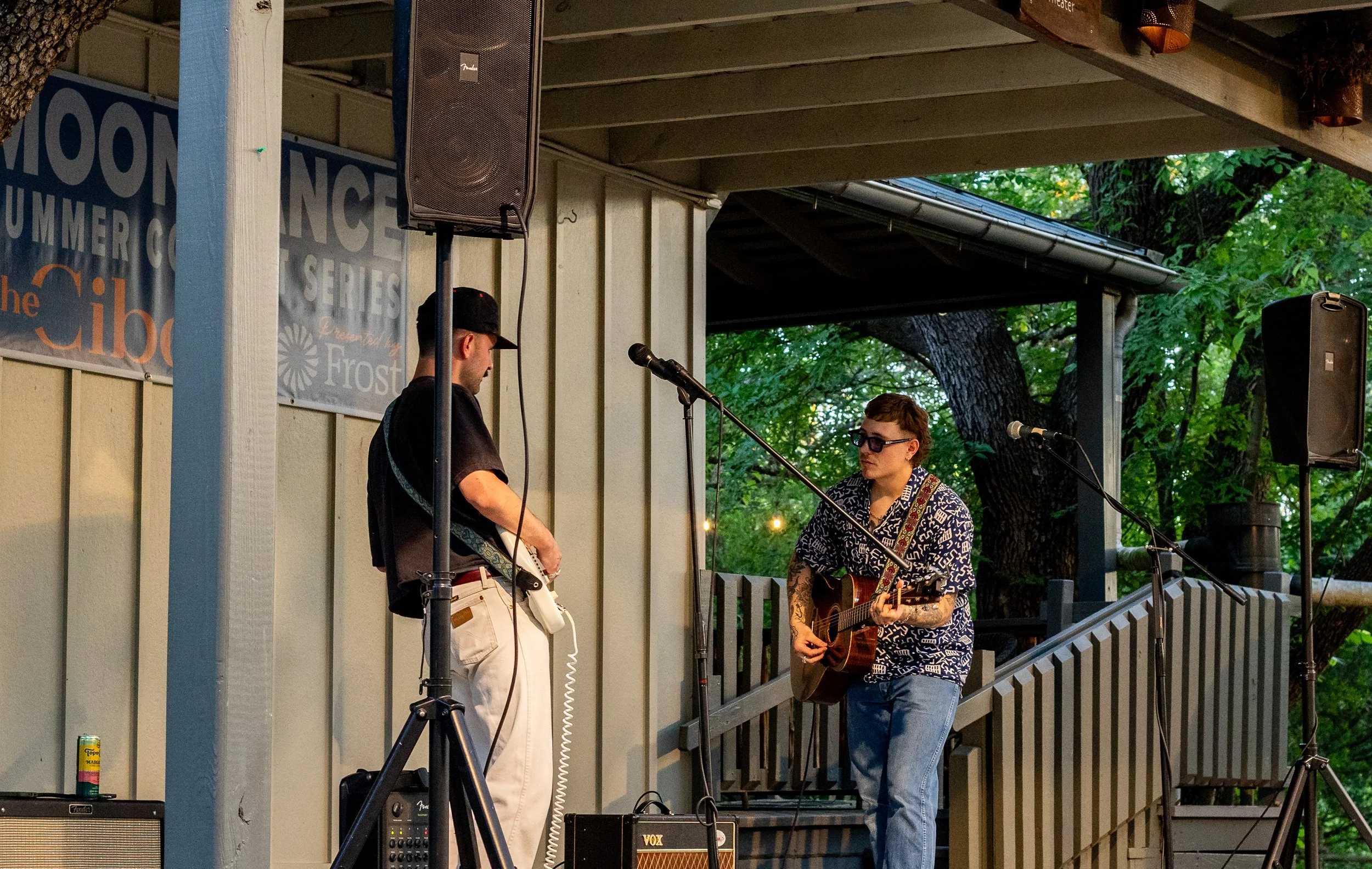 Two musicians performing on a small outdoor stage surrounded by trees. One is playing an electric guitar, and the other is playing an acoustic guitar and singing into a microphone. There are large speakers and musical equipment on the stage.