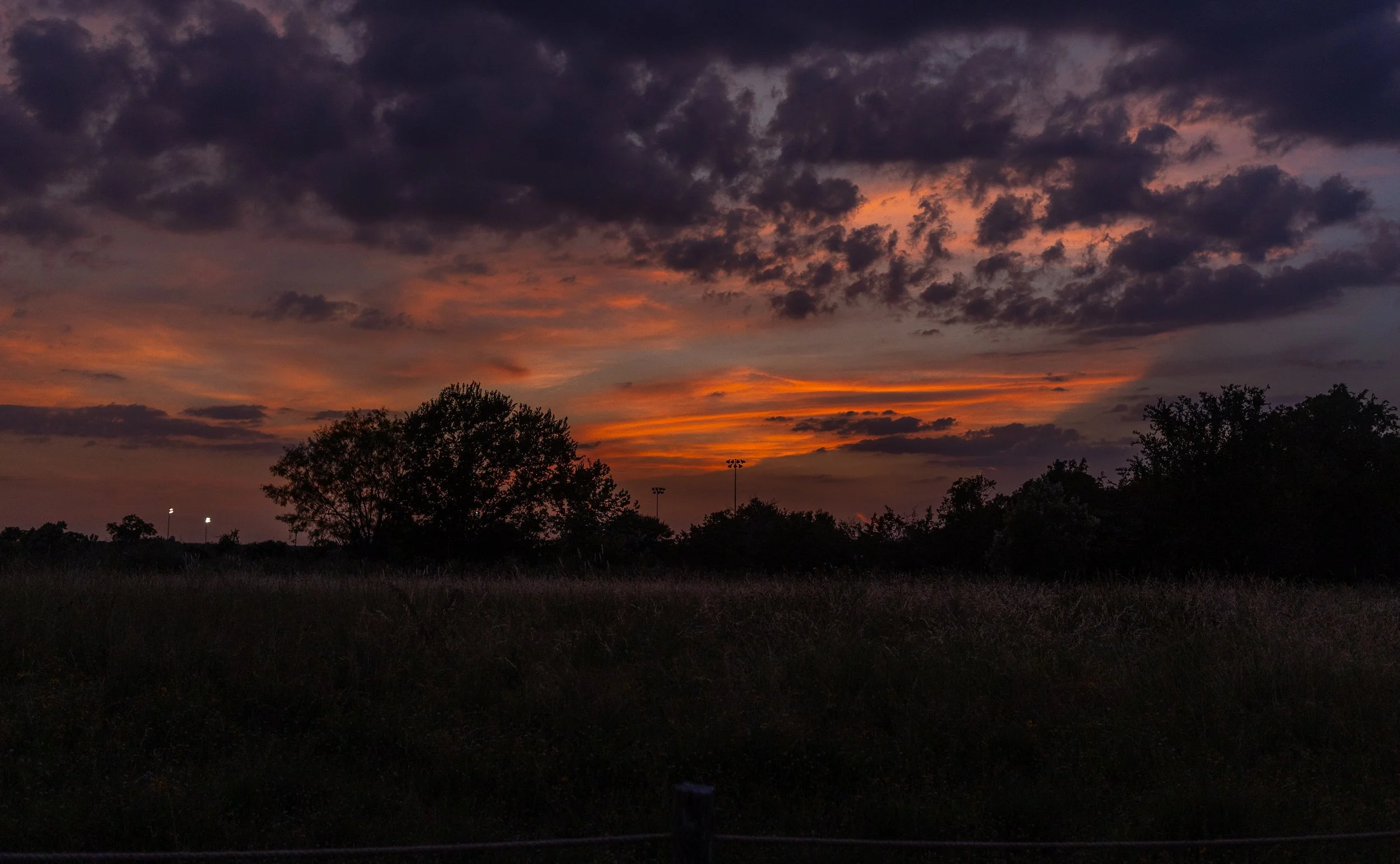 A sunset sky filled with dark and orange clouds over a silhouette of trees and sports field lights.
