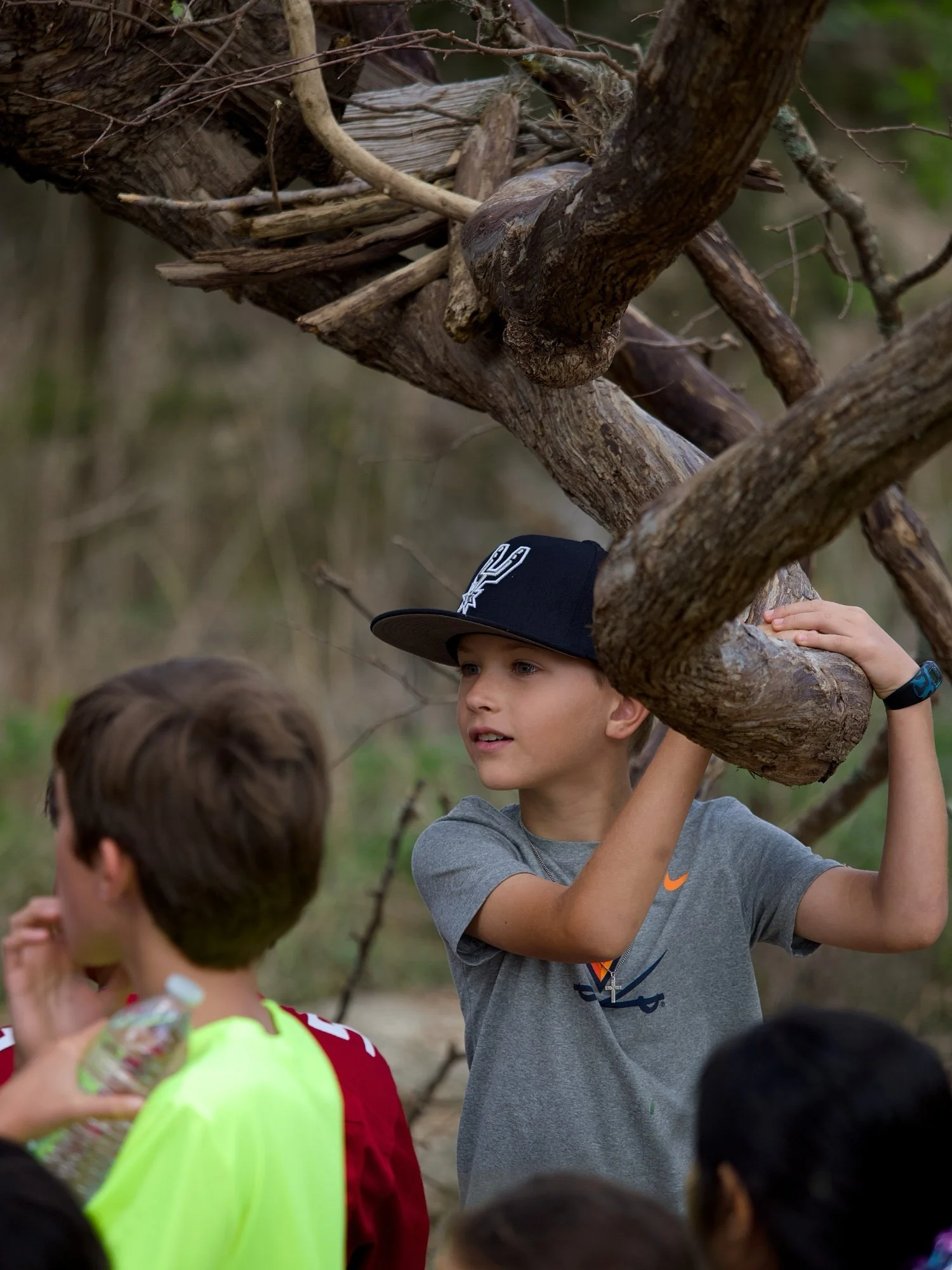 A group of children outdoors, gathered around a tree with thick branches. One boy in a gray Nike t-shirt and a black baseball cap is holding onto a branch, while others look on, some holding drinks or other items. The background appears to be a natur