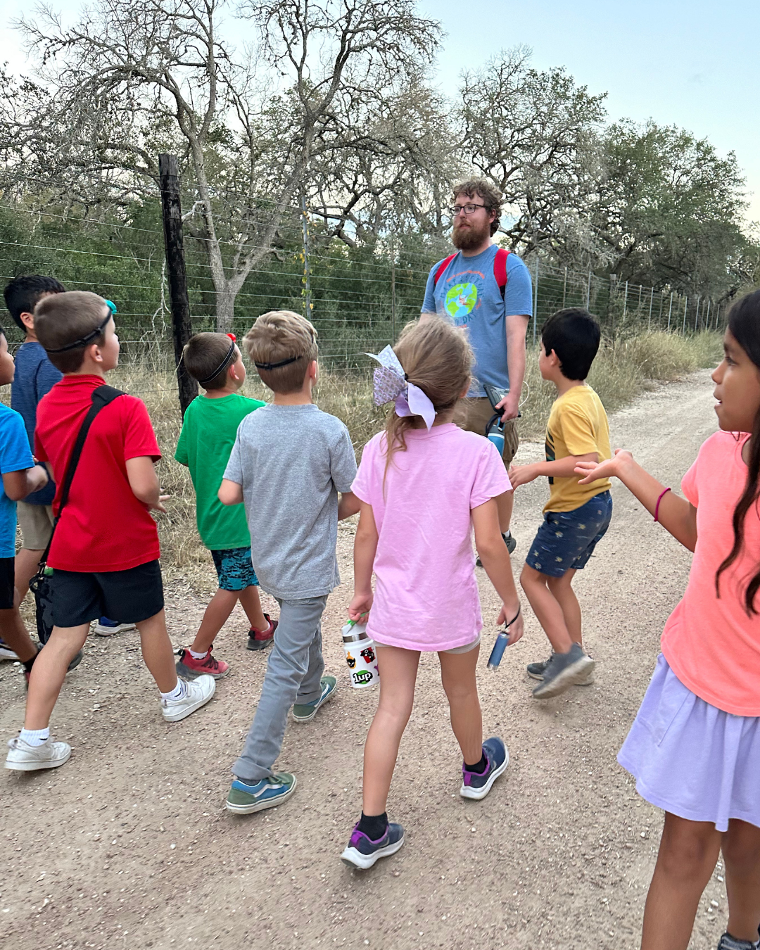 A man with a beard and glasses, wearing a blue t-shirt and carrying a red backpack, standing in front of a group of children on a dirt path outdoors, with trees and a fence in the background.