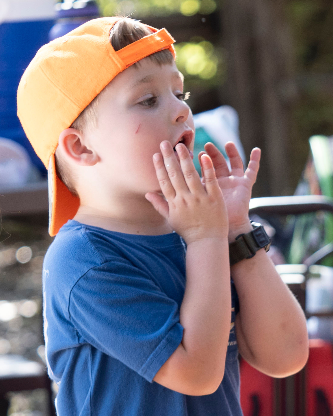 A young boy wearing an orange baseball cap backwards and a blue t-shirt is singing a cheer. The background is outdoors with some blurred objects and trees.