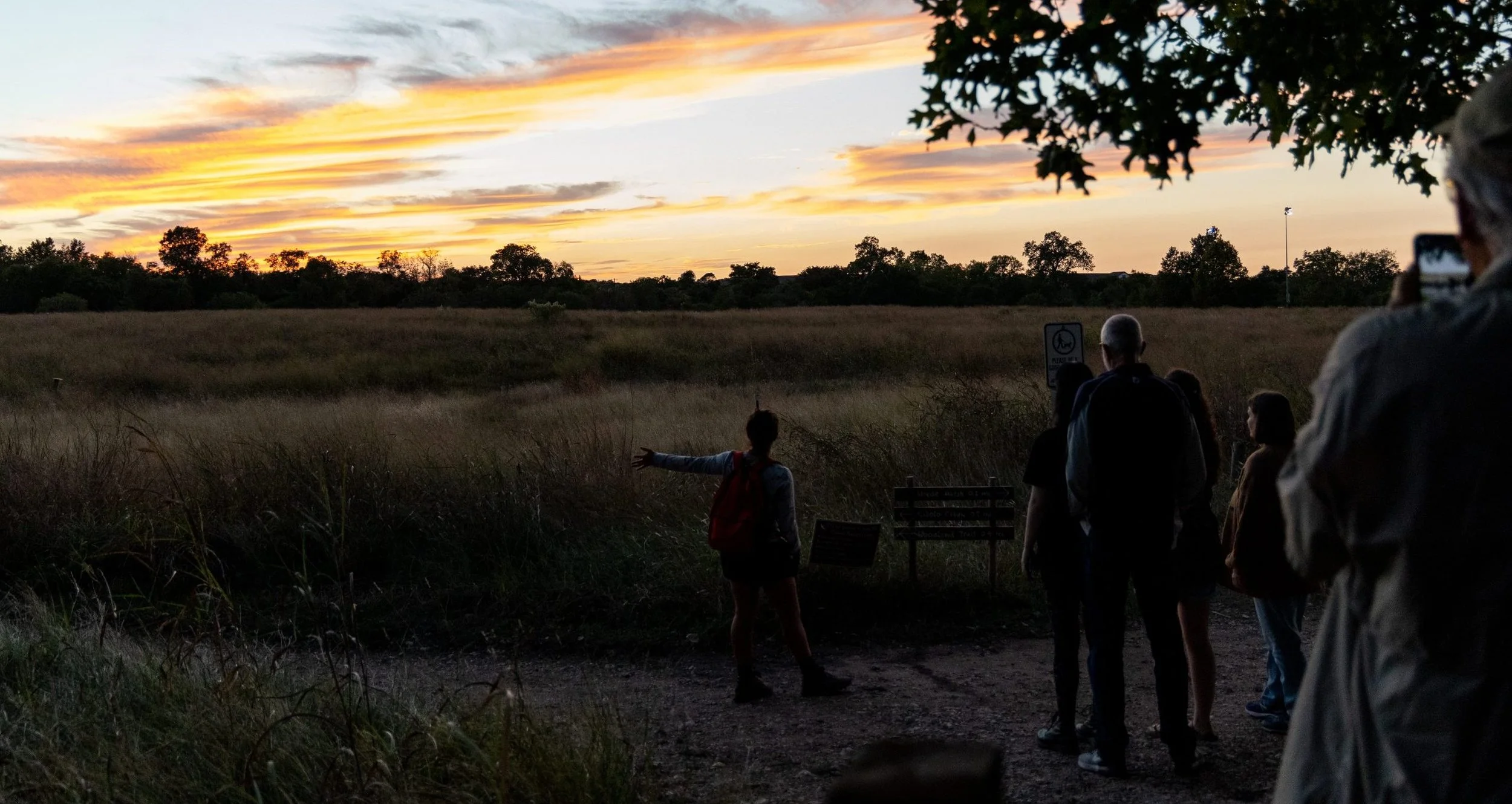 Group of people watching a sunset on a night hike at the cibolo, with some pointing towards the sky and others taking photos. The sky has orange and pink clouds, and there are trees and open fields around.