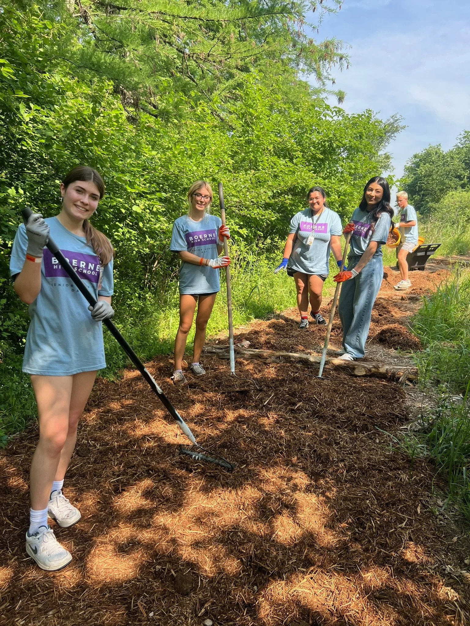 A group of five young people, four women and one man, engaged in a gardening or outdoor activity along a wooded trail. They are holding gardening tools and wearing casual clothes with gloves, smiling at the camera.