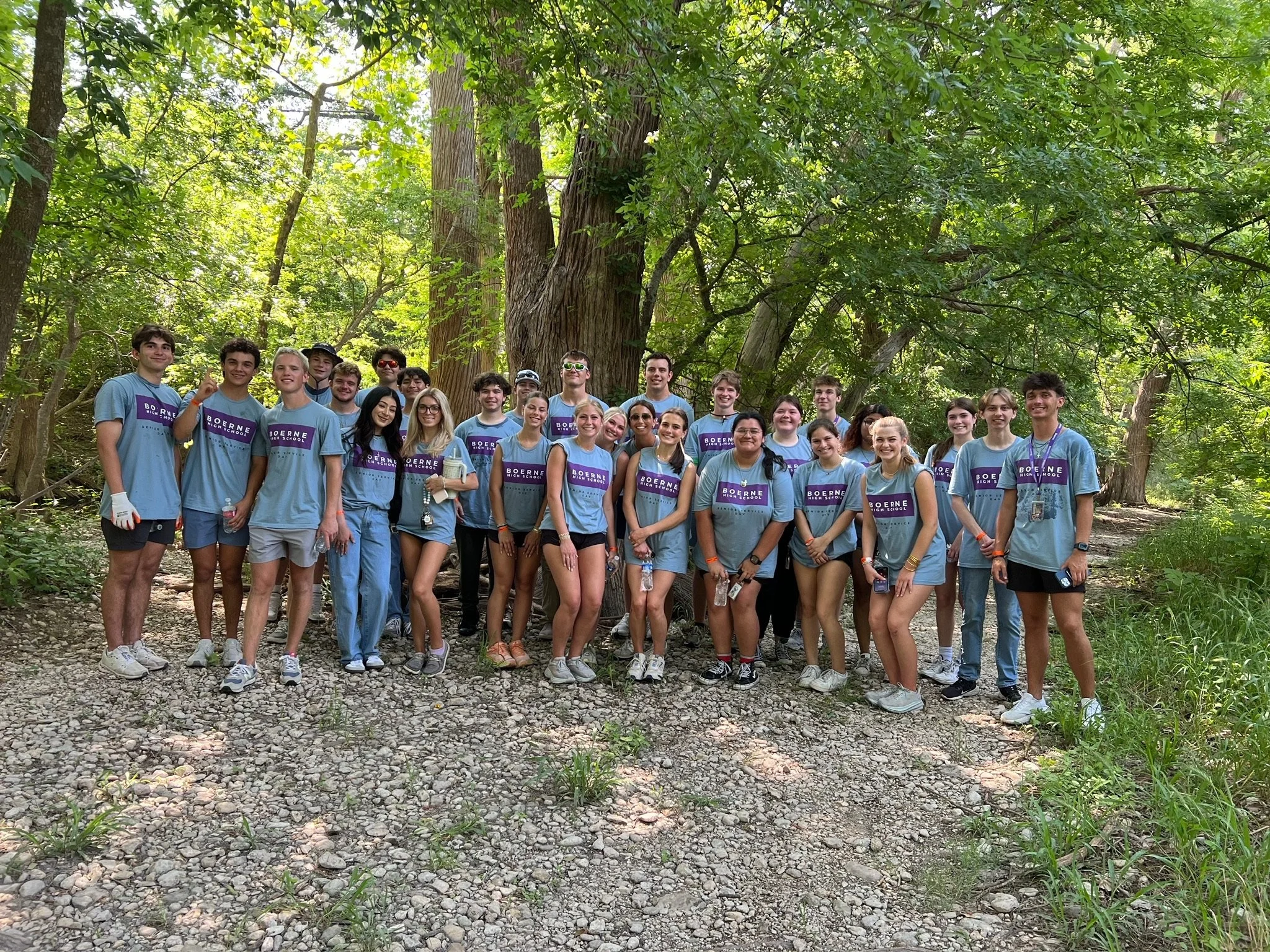 A group of young people in matching blue t-shirts with 'Boerne High School' written on them, standing together outdoors in a wooded area with tall trees and sunlight filtering through the leaves.