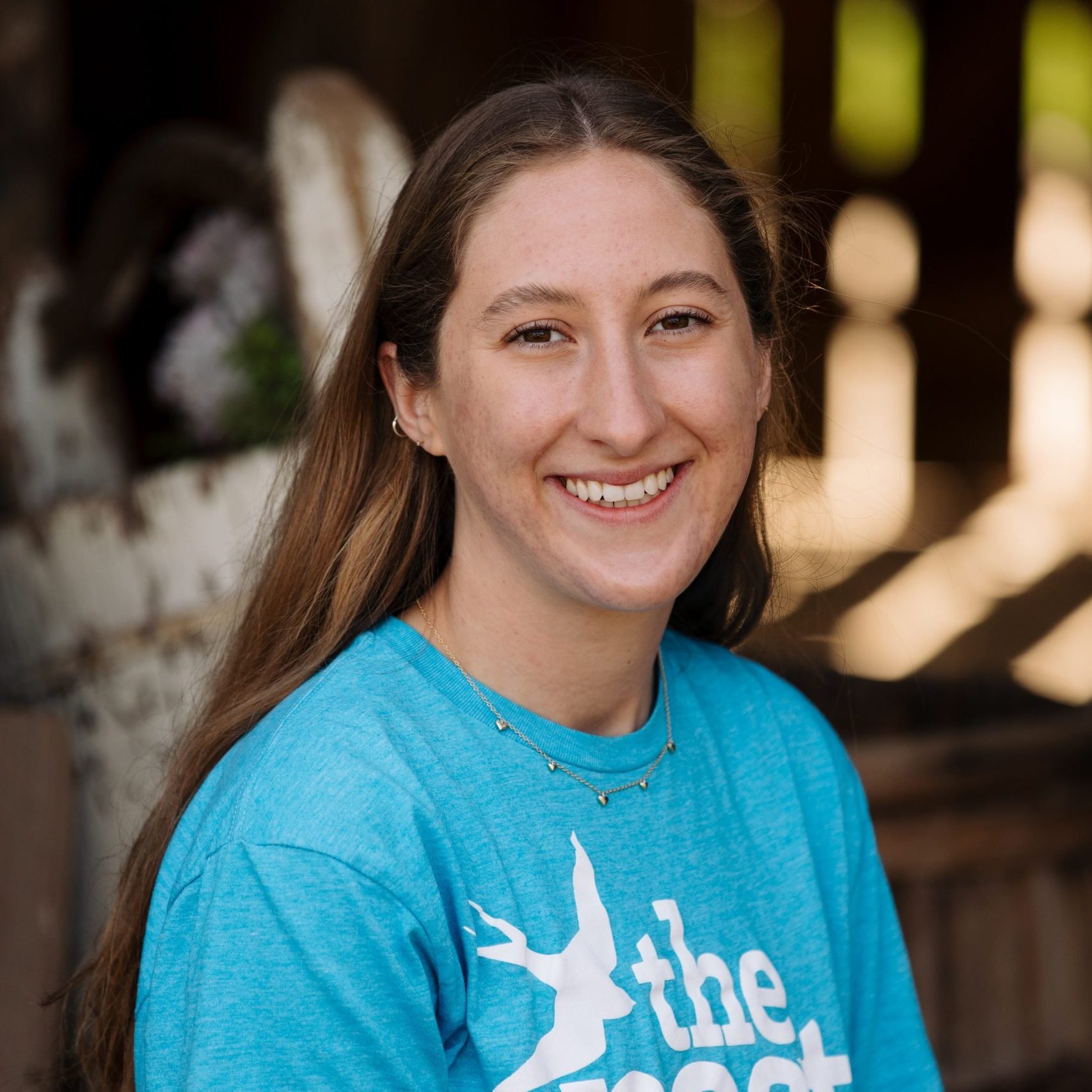 A young woman with long brown hair, smiling, wearing a bright blue T-shirt with white text, a delicate necklace, and small hoop earrings. She is standing outdoors with blurred wooden fence and greenery in the background.