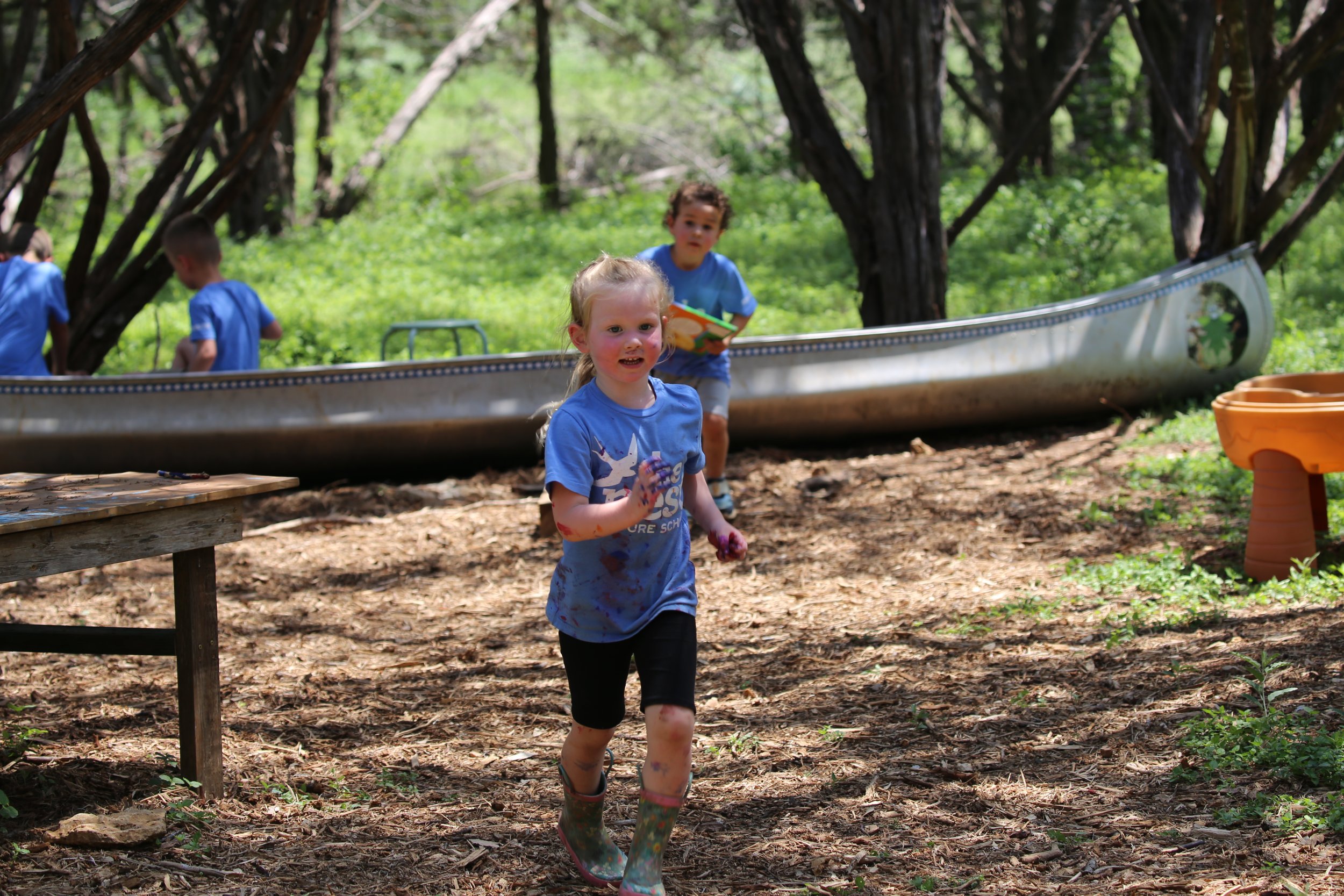 Children playing outdoors in a wooded area, with one girl running toward the camera and others in the background near a canoe.