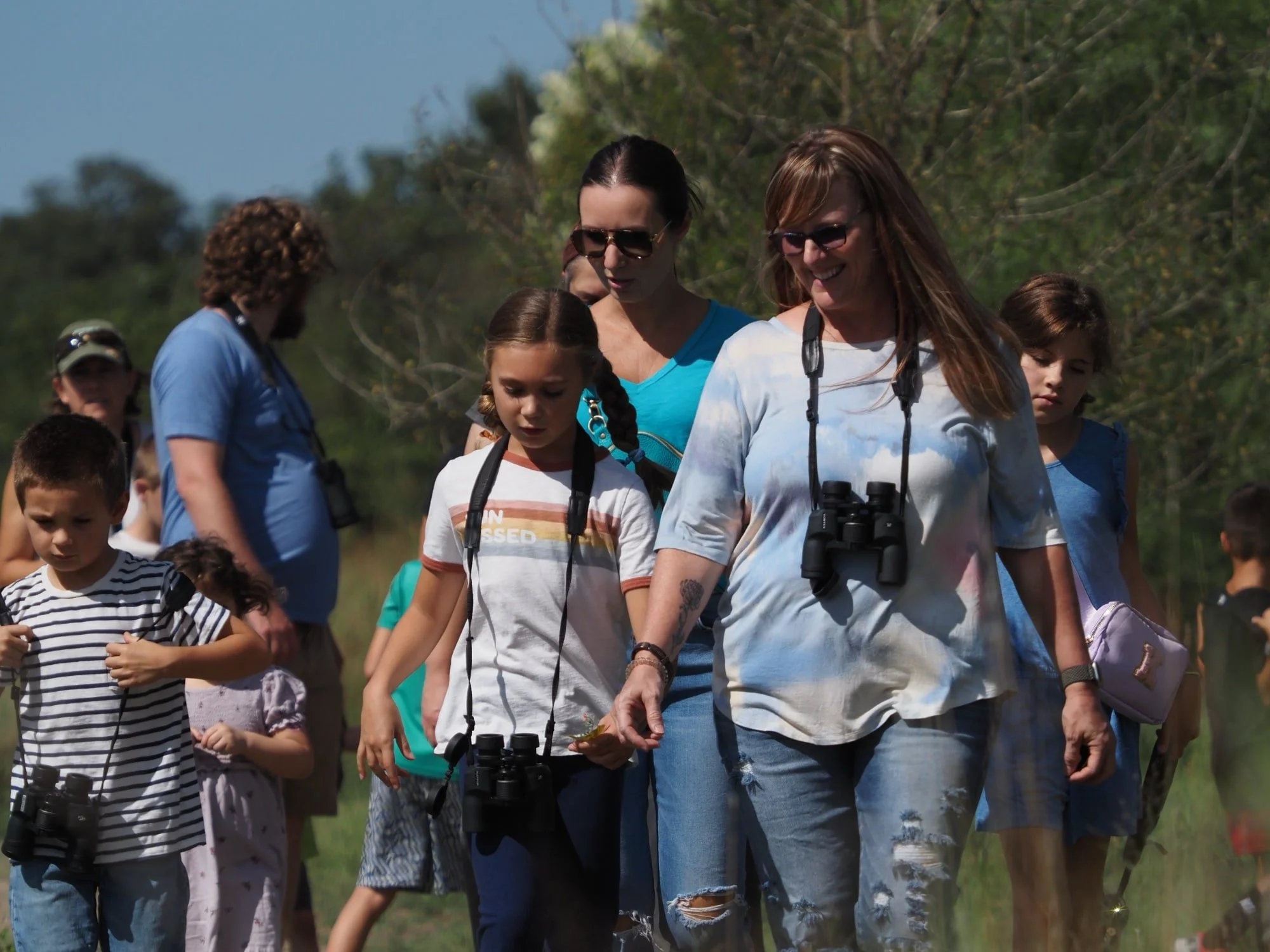 Group of children and women walking outdoors, some holding binoculars, under a clear blue sky with greenery in the background.