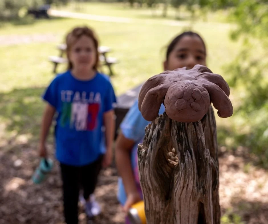 Two children outdoors, with a clay sculpture of a spider in focus in the foreground.