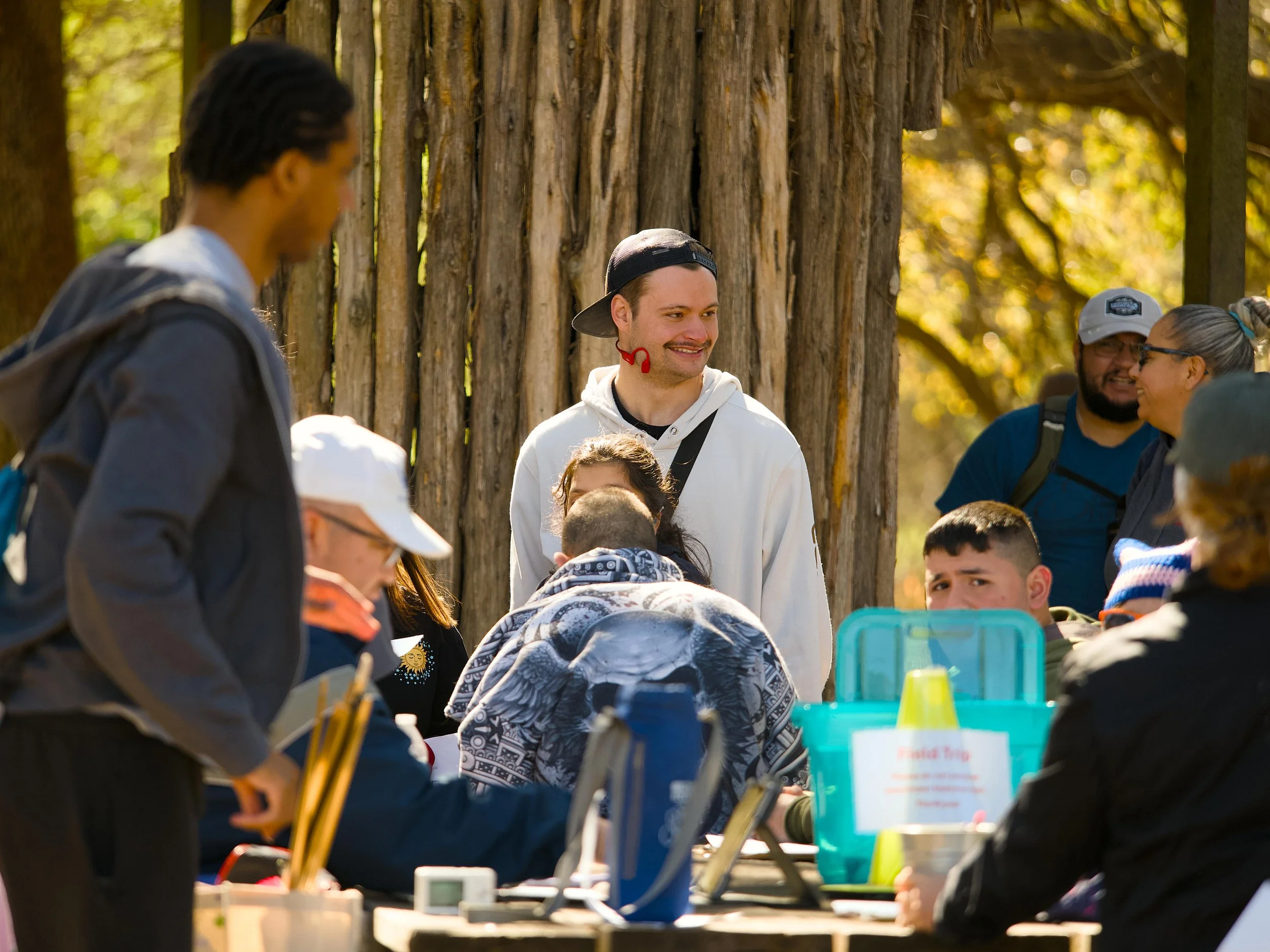 Group of people, including children and adults, gathered outdoors around a table with various objects, with a large wooden fence and trees in the background, on a sunny day.