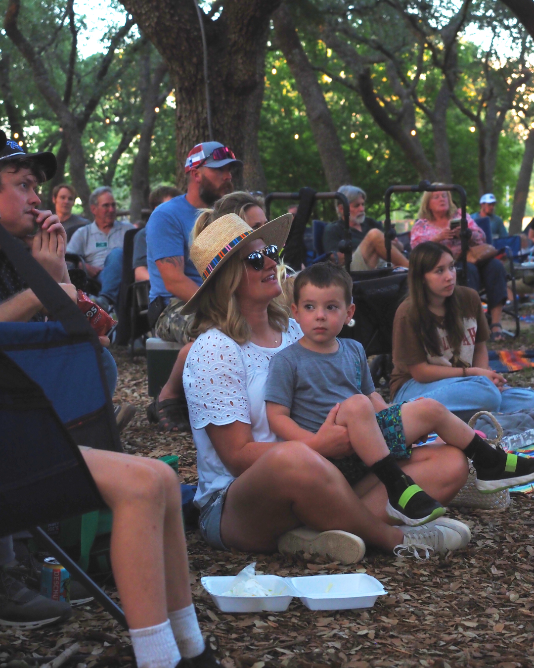 People sitting outdoors in a wooded area, watching a performance or event during the evening. The group includes a woman with a straw hat and sunglasses holding a young boy on her lap, and several others seated on the ground or on chairs, with trees 