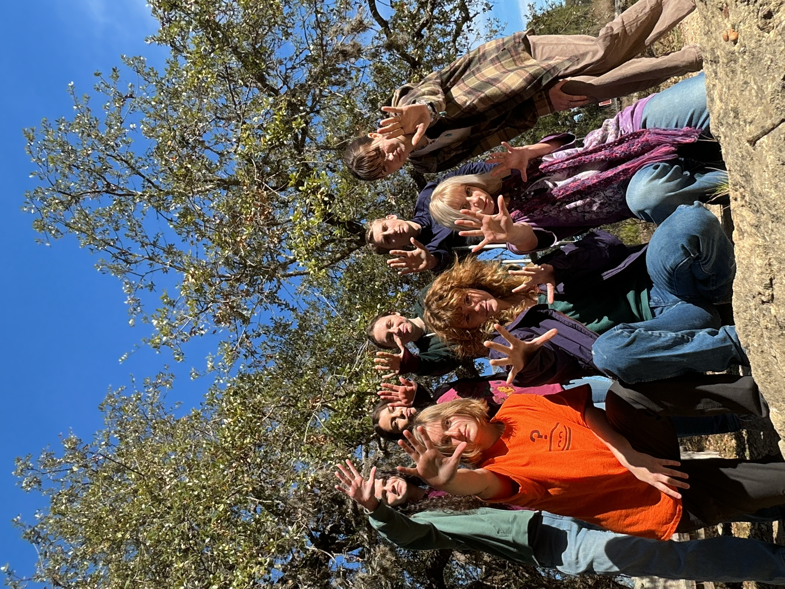A group of diverse people posing outdoors under a tree on a sunny day, making peace signs and smiling.