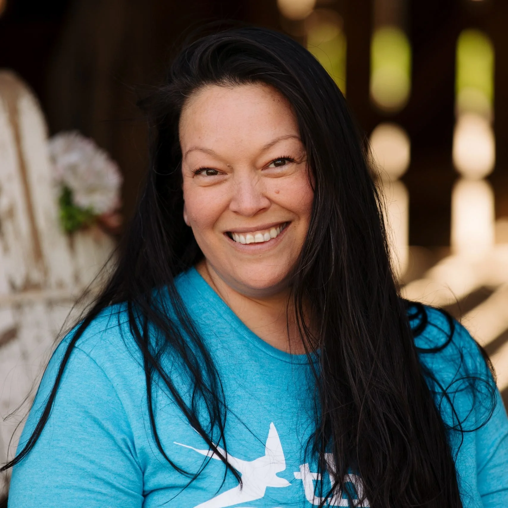 Close-up of a smiling woman with long black hair wearing a blue shirt with a white design, standing outdoors near a wooden structure with blurred background.