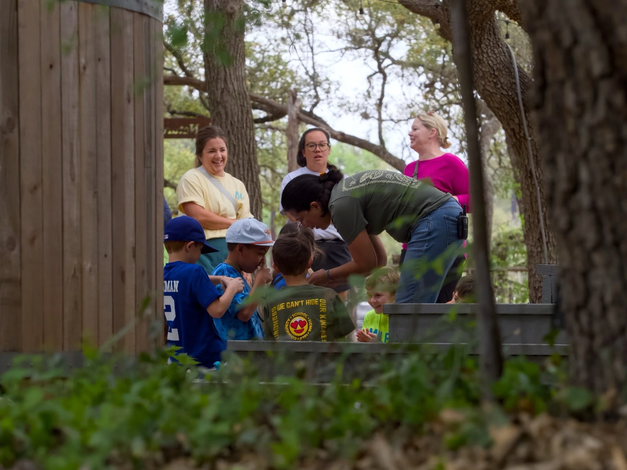 Children and adults gathered outdoors, involved in an activity, with trees and a wooden fence in the background. Some children wear blue shirts and white hats, while adults are dressed casually, one in a pink top and another with glasses and a bun. T
