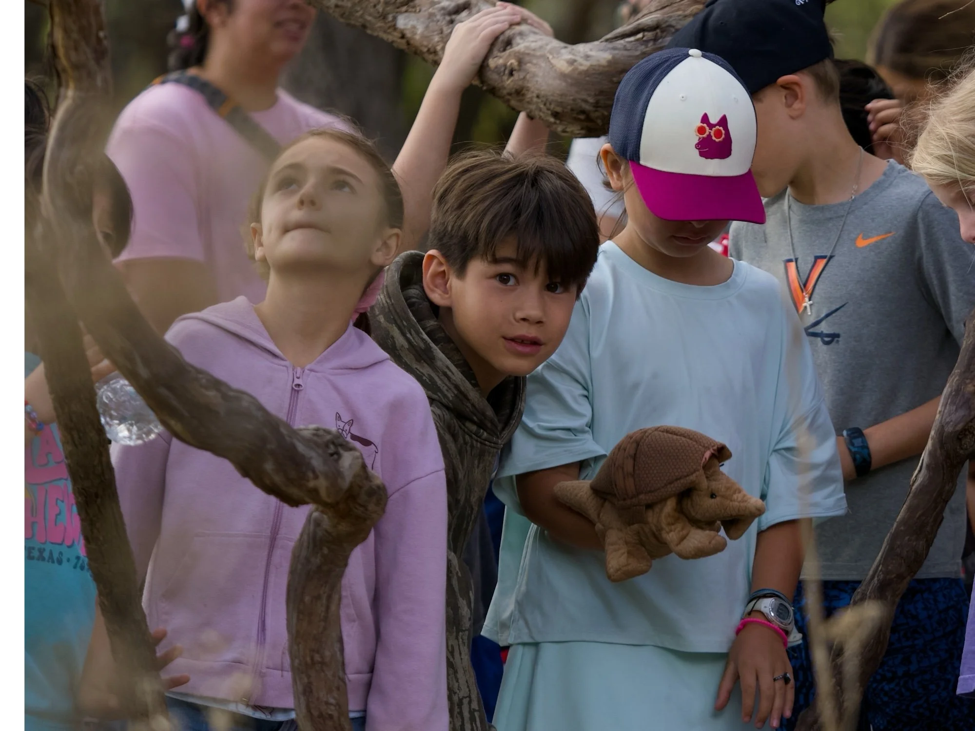 Group of children outdoors, some holding a bear-shaped costume, with trees and blurred faces in the background.