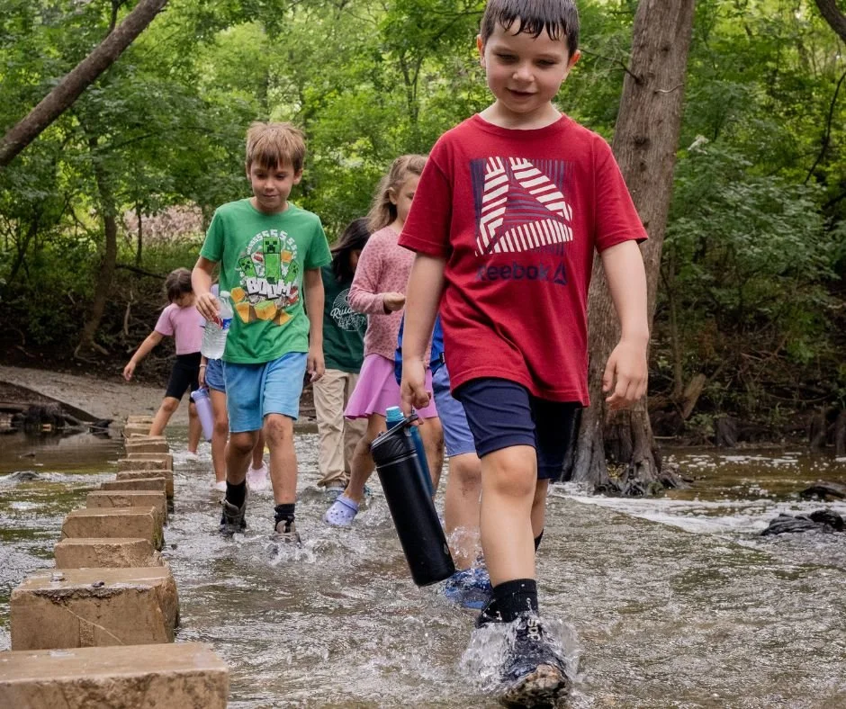Children crossing a shallow Cibolo Creek in a wooded area, some carrying water bottles, walking on stepping stones.