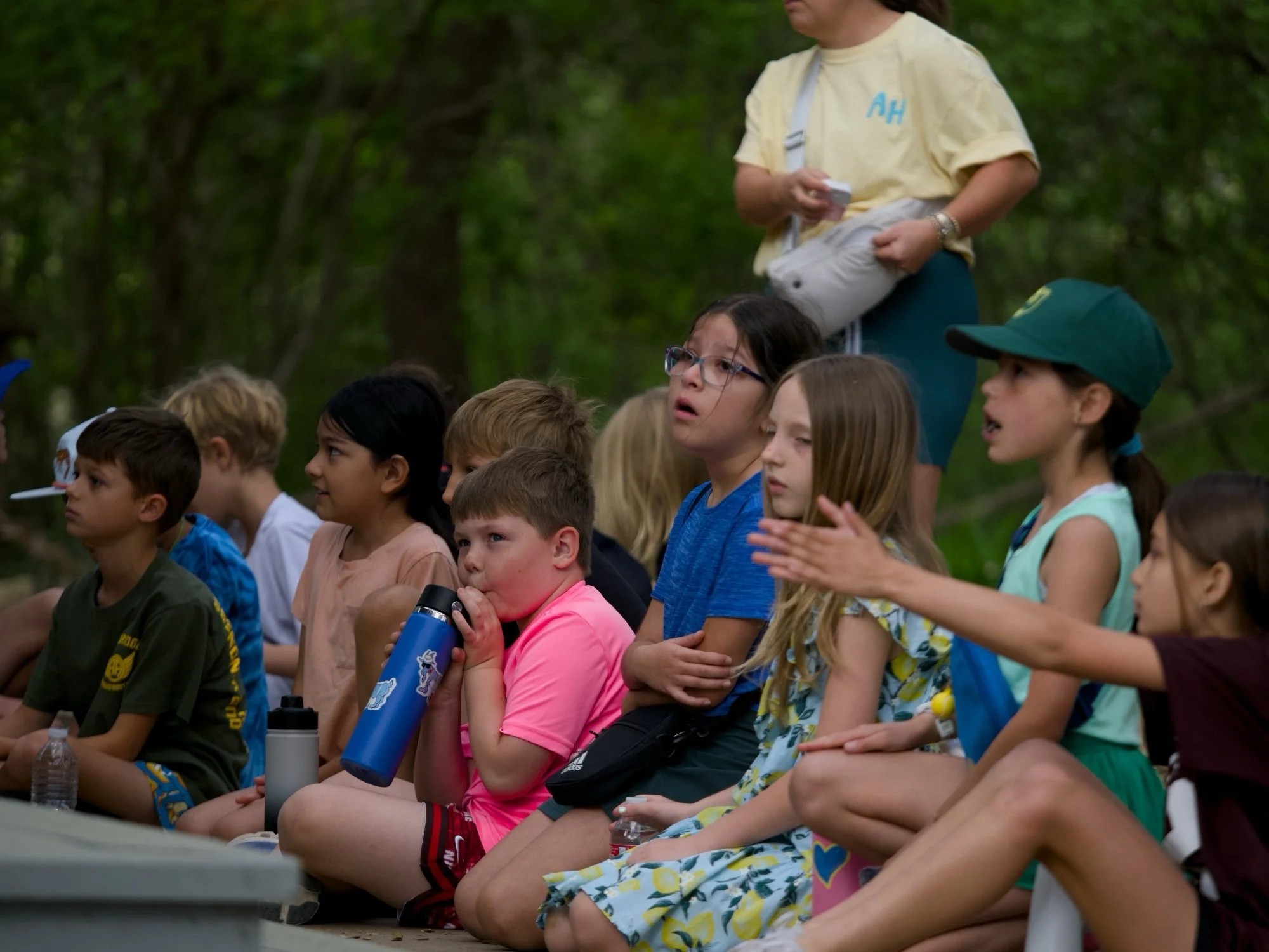 A group of children sitting outdoors on a sunny day, attentively listening, some holding water bottles or cups, with trees in the background.
