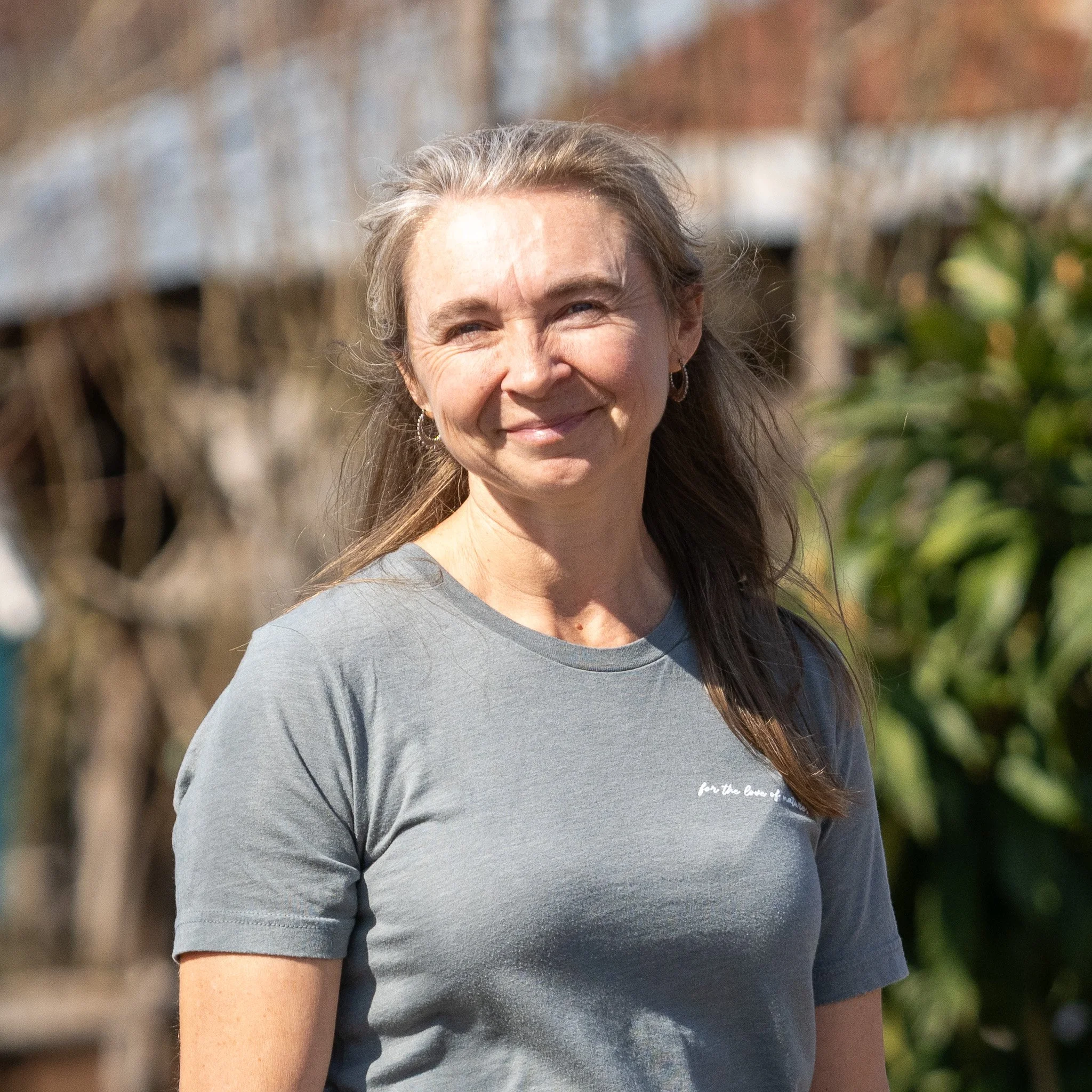 A woman with grayish-brown hair, smiling, wearing a gray t-shirt with small white text on it, standing outdoors with trees and a wooden fence in the background.