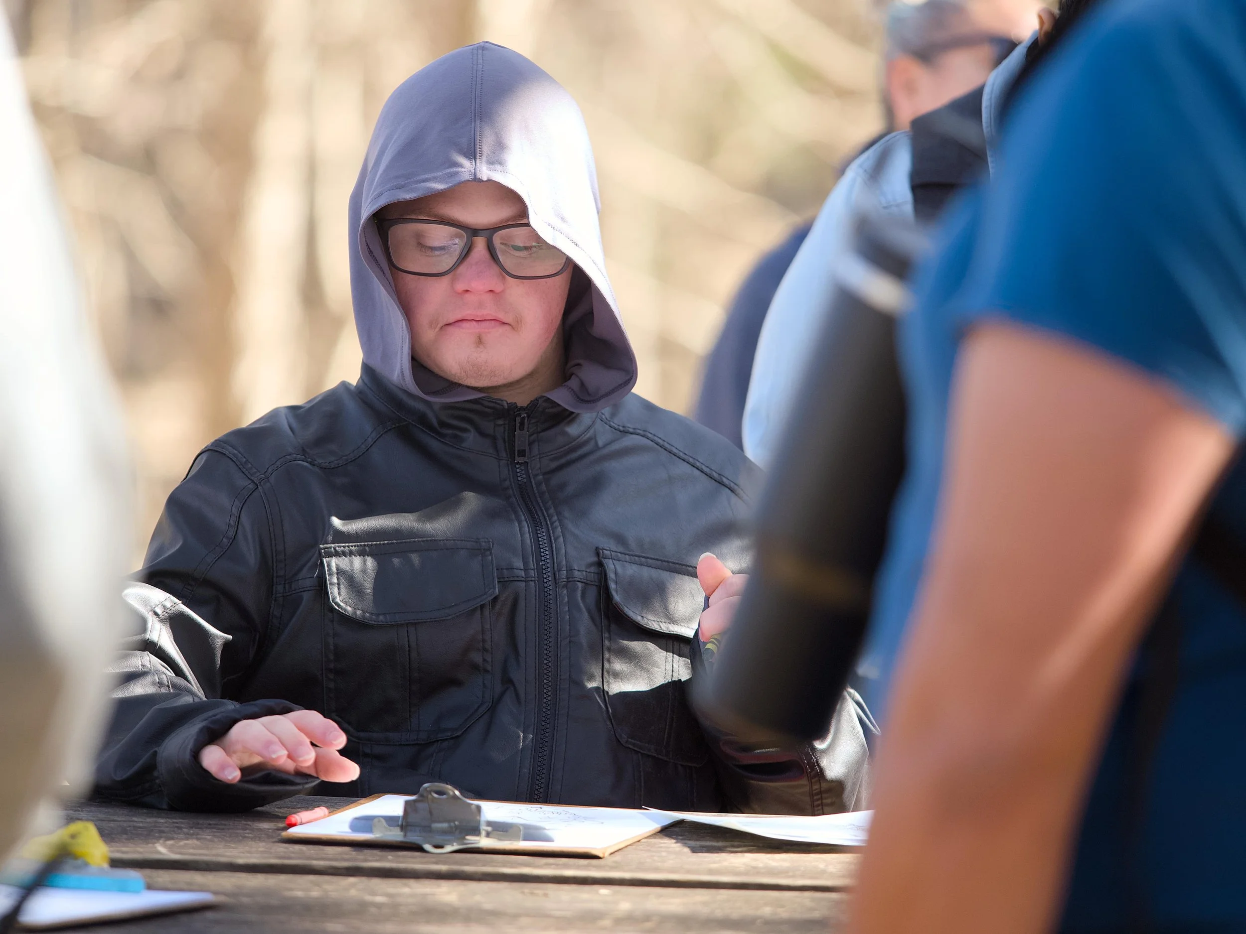 A person with glasses wearing a hoodie and leather jacket is looking down at a table with papers and a clipboard, with others partially visible around him outdoors.