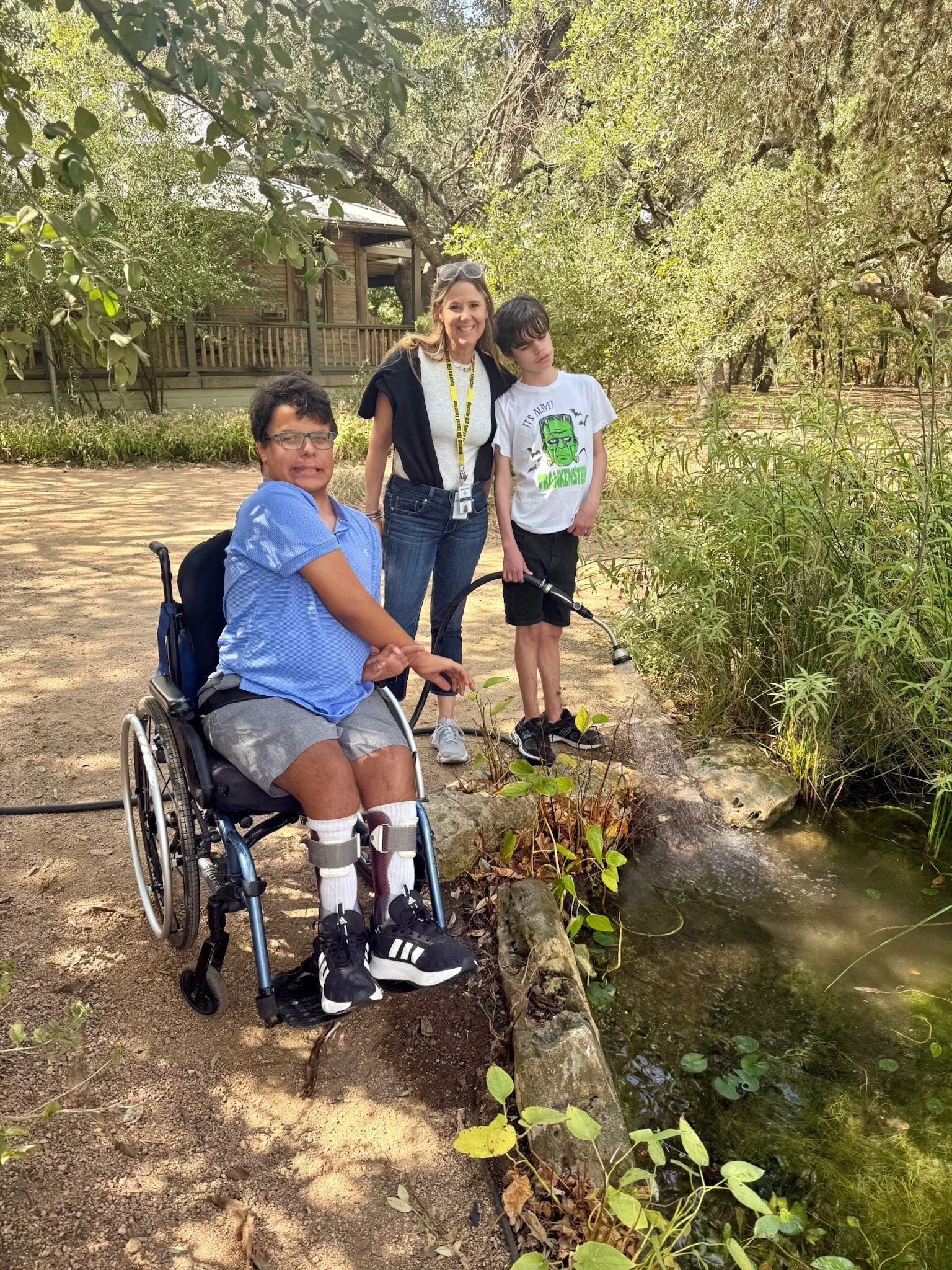Three people outdoors near a small creek, with trees and a wooden house in the background. One person is in a wheelchair, two others stand beside the creek, one holding a water hose. They are smiling or looking at the water.