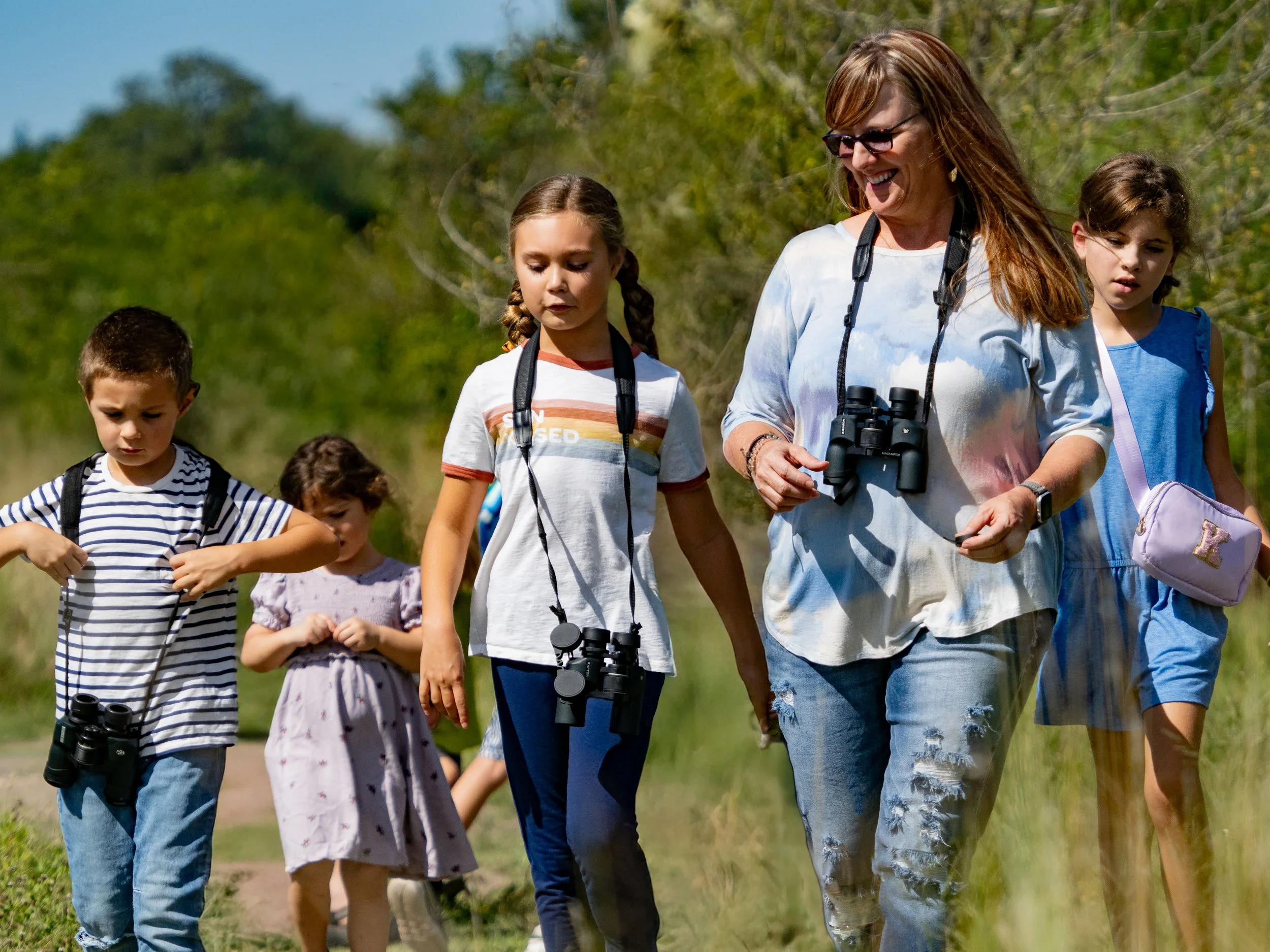 A woman and several children walking outdoors on a nature trail carrying binoculars, with trees and blue sky in the background.