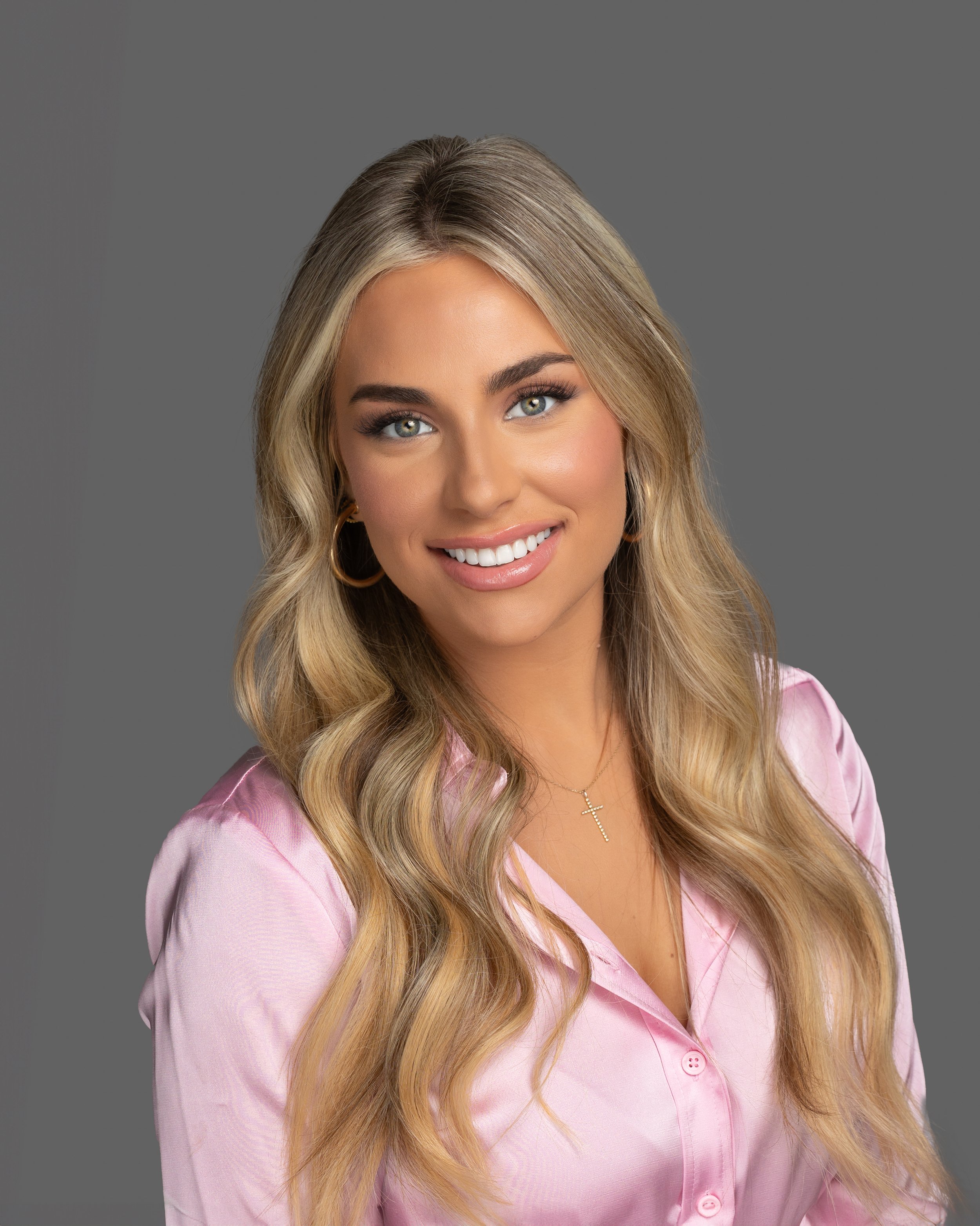A young woman with long, wavy blonde hair and light skin, smiling, wearing a pink satin blouse, gold hoop earrings, a delicate gold cross necklace, and makeup, against a plain gray background.
