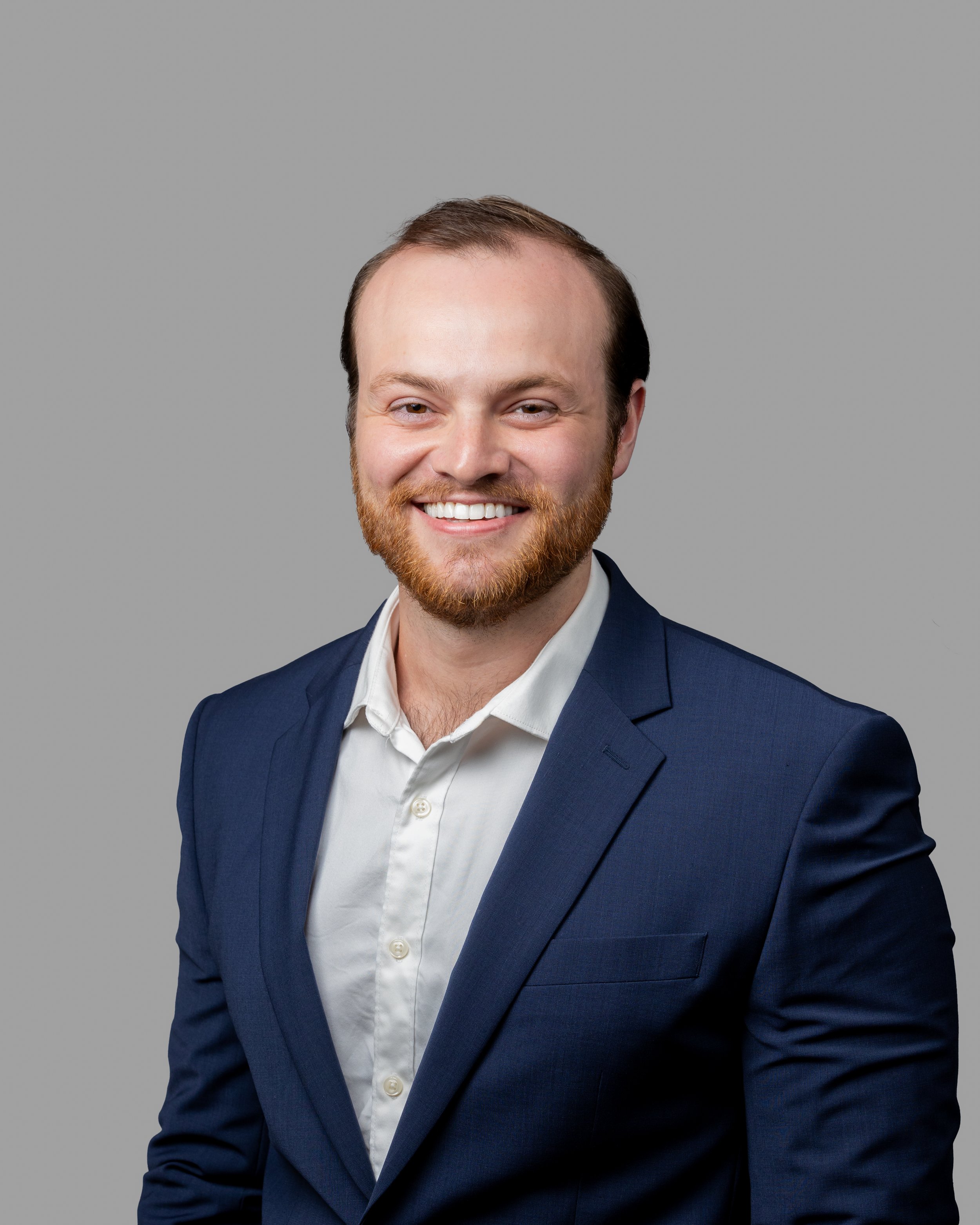 Headshot of a smiling man with a beard and short hair, wearing a navy blue suit and white shirt, against a plain gray background.